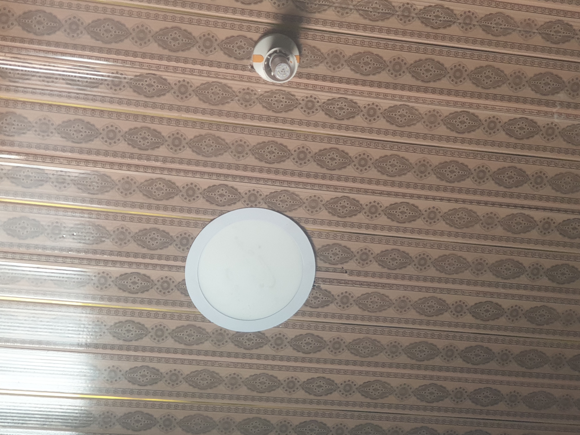 An interior shot captures a patterned ceiling with two light fixtures. A circular white LED panel light is positioned on the lower left, while a smaller, exposed bulb fixture with an orange accent is located on the upper center. The ceiling itself is covered in a repeating brown and tan geometric pattern.