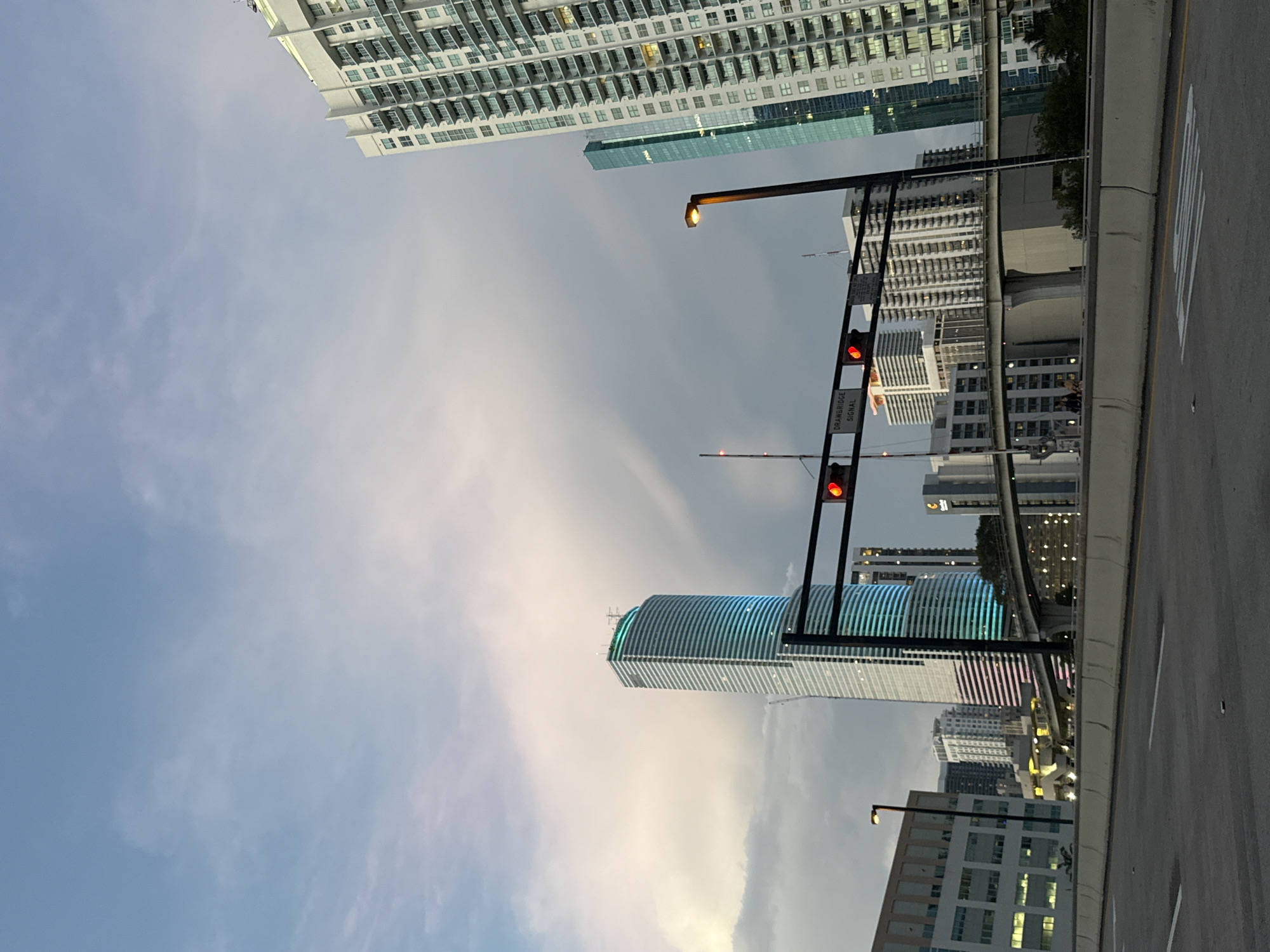 The image shows a street scene in Miami, Florida, USA. The view is looking upwards at tall buildings, including one with a distinctive curved, striped facade. A traffic light pole with red lights is visible in the foreground. There is a street and some buildings. The sky is partly cloudy.