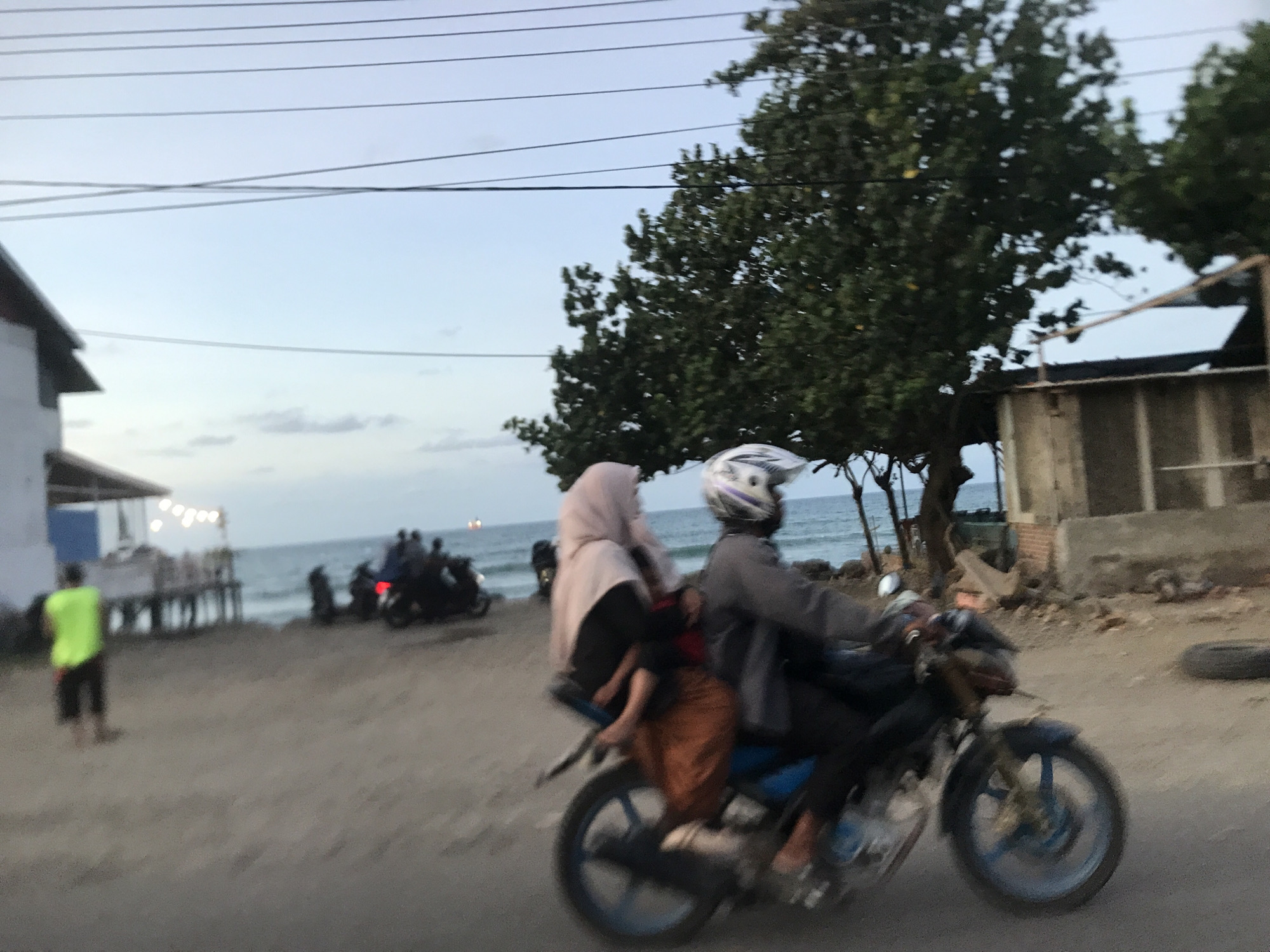 The image, likely taken at dusk in the city of Lhokseumawe, Indonesia, presents a bustling coastal scene. In the foreground, a blue motorcycle is in motion, carrying a rider wearing a helmet and a passenger in a hijab, who holds a child in her lap. To the left, a person in a neon yellow shirt stands near a beachside structure with string lights. In the background, several people and motorcycles are gathered near the shoreline. Beyond them is the ocean, with faint horizontal clouds near the horizon. To the right, there is a tree and a partially constructed or dilapidated structure made of cement or concrete. Wires are visible above the scene. The ground appears to be a dirt or gravel road. A spare tire is visible to the right.