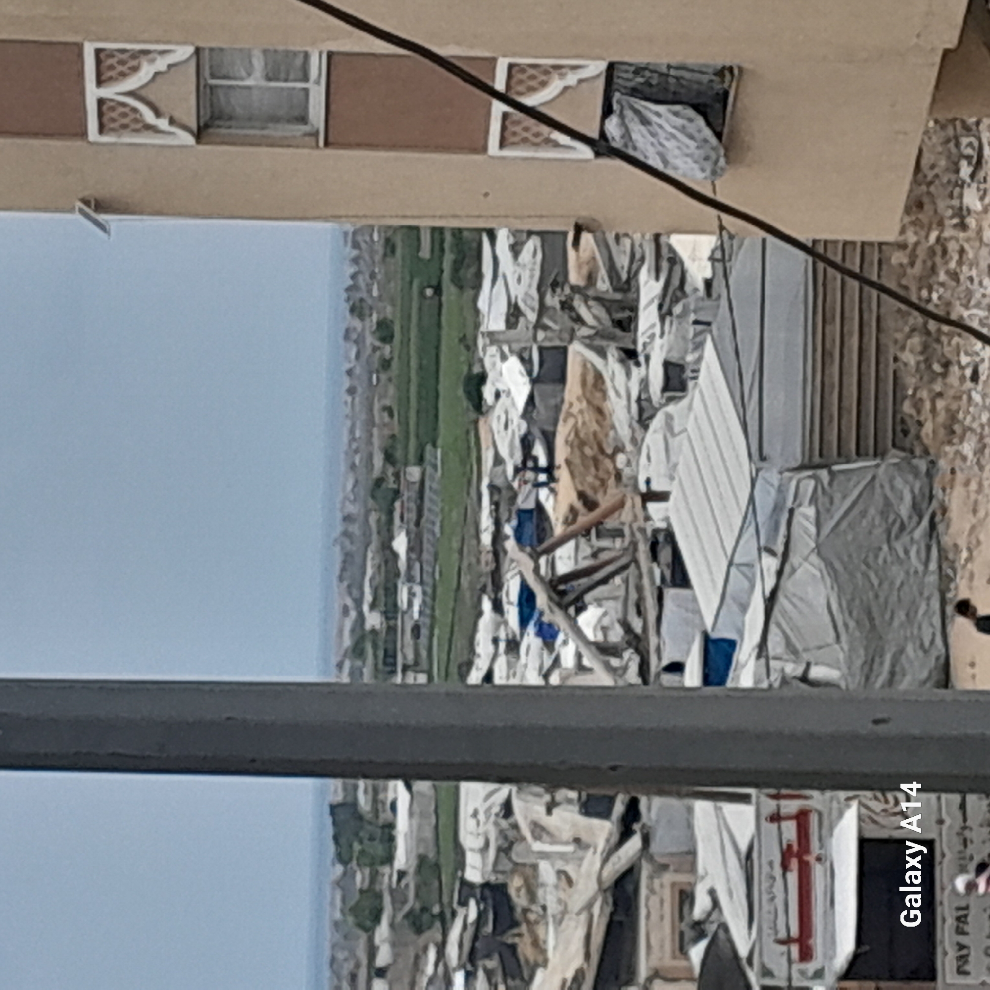 A photograph captures a panoramic view of what appears to be an informal settlement or refugee camp, likely situated in a developing area or a region afflicted by displacement. The camp is characterized by a dense and haphazard arrangement of temporary shelters, primarily constructed from white and gray tarpaulins and scavenged materials. The structures are closely packed together, creating a sense of overcrowding and limited space. The surrounding landscape consists of fields and a distant body of water. The image is taken from an elevated perspective, possibly from a building or high ground overlooking the camp. The foreground features parts of a building's architecture, including windows with decorative features and a steel beam, suggesting the photograph was taken from an inhabited structure. In the lower right corner of the image, the text "Galaxy A14" is visible, indicating the device used to capture the photo. The overall tone of the image is somber, highlighting the challenging living conditions and the precariousness of the settlement. The location is visually described as a densely populated informal settlement in a region with surrounding agricultural land.
