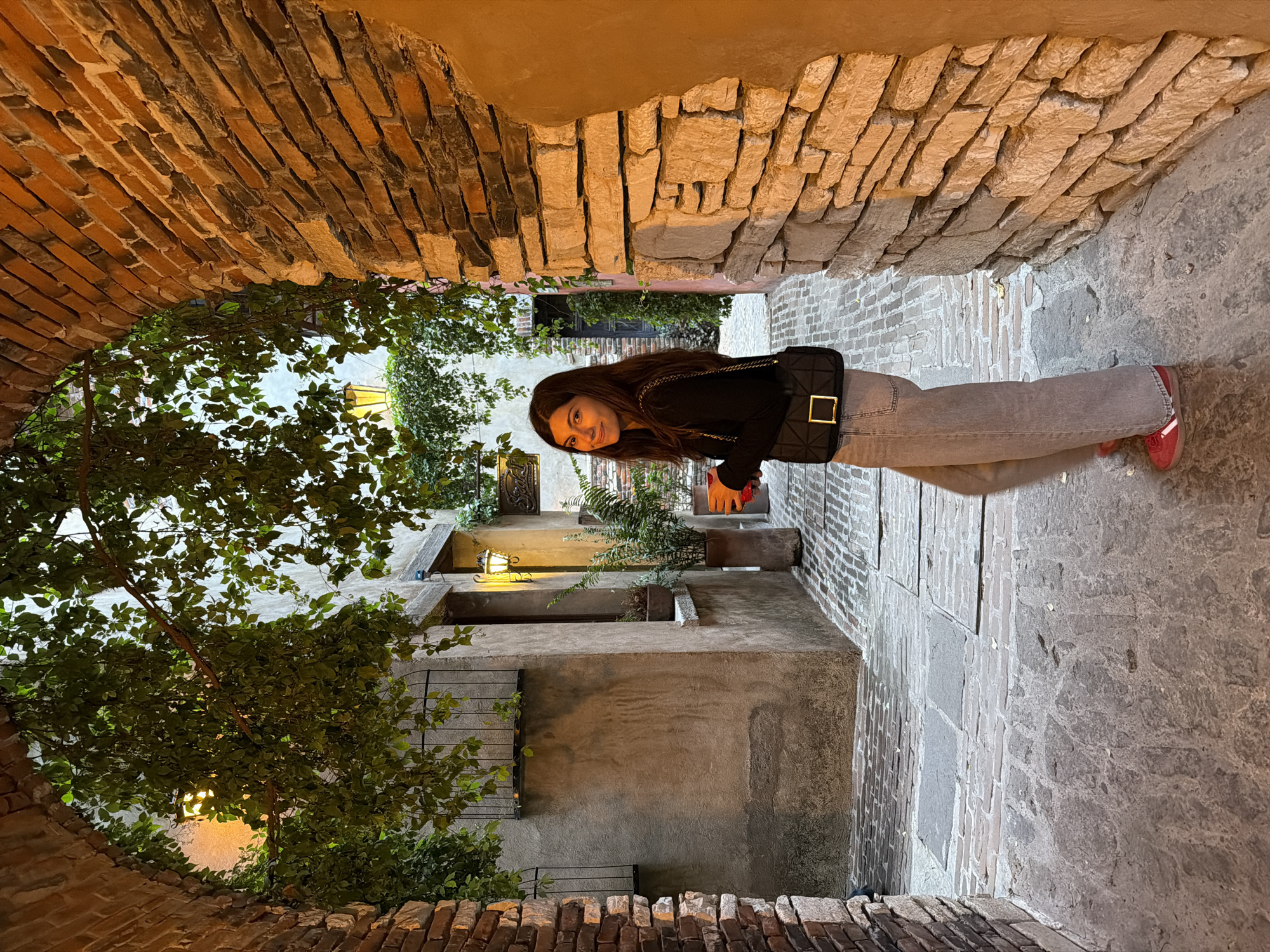 A young woman stands in a narrow alleyway in Domingo Arenas, Mexico. The alley is enclosed by brick walls and an arched brick ceiling. She wears a black long-sleeve shirt, light gray jeans, and red sneakers, and carries a black quilted bag. The alley floor is paved with stone. A tree with dense foliage is on the left side of the frame. In the background, a building with plants and a lit lantern is visible. The lighting is warm and subdued, suggesting the photo was taken in the evening.