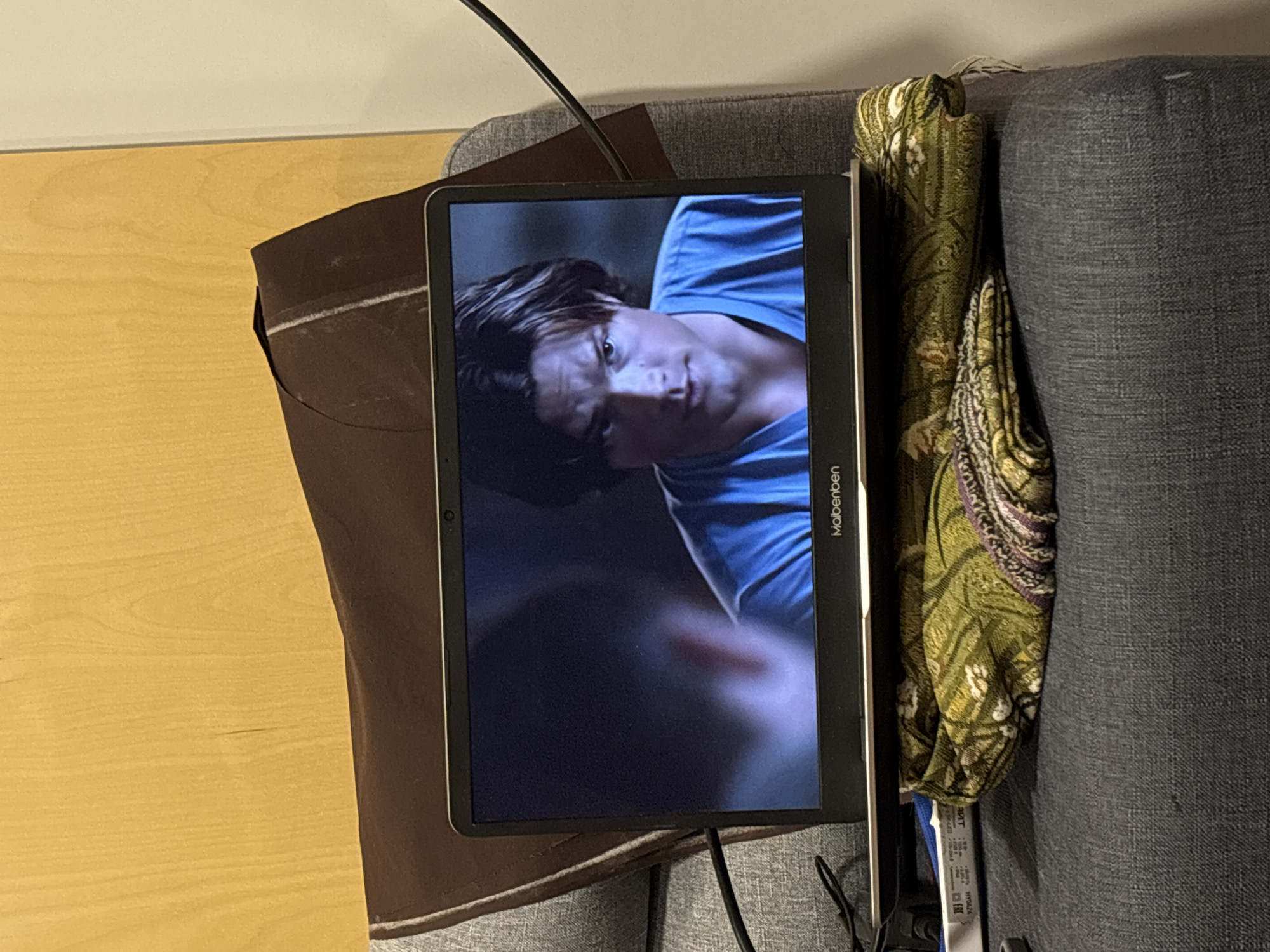 A tablet is sitting on a grey couch. The screen shows a person with dark hair and a blue shirt. A brown cloth is to the left of the tablet, and a green patterned cloth is to the right. The word "Malbenben" is on the side of the tablet. A black cable is connected to the tablet. A beige-colored surface is partially visible in the upper-left corner. The location in Perm, Russia is unknown.