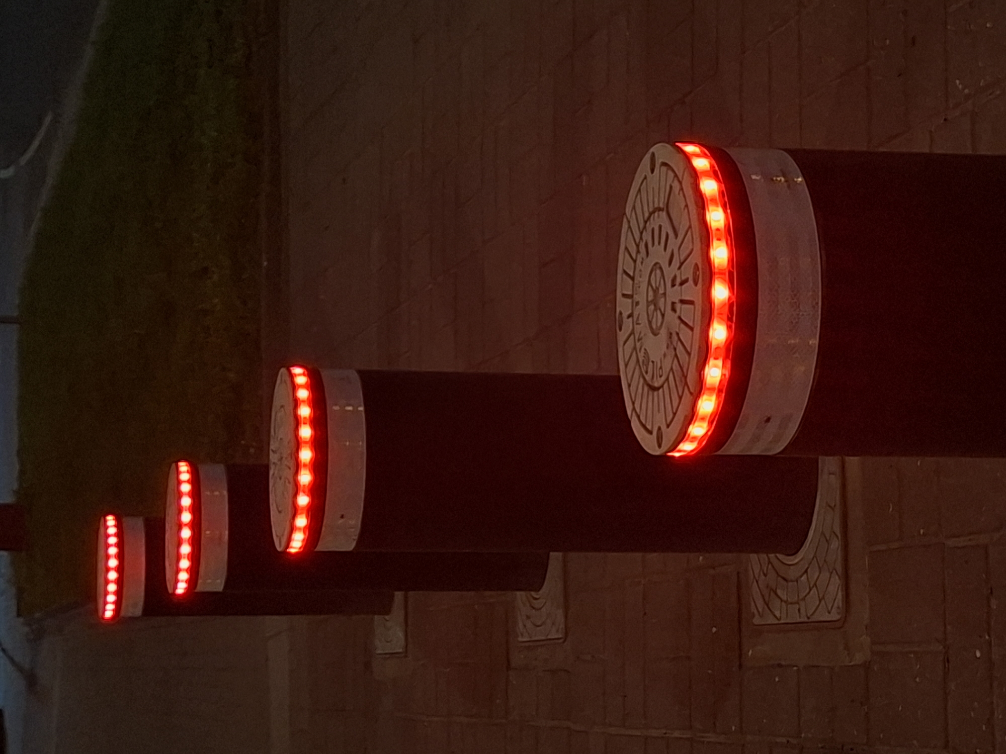 The image shows a row of bollards with red lights along a paved pathway. The bollards are cylindrical and dark in color, with a white band below the illuminated tops. The pathway is made of interlocking brick-like paving stones. In the background, there is a grassy embankment and a road. The scene appears to be at dusk or at night, as indicated by the illuminated bollards and the dim lighting. The location is specified as Tallinn, Estonia. There are no people or animals visible in the image. The bollards are spaced evenly and arranged diagonally from the foreground to the background, receding into the distance. The top of the bollard in the foreground has a circular metal plate with embossed patterns.