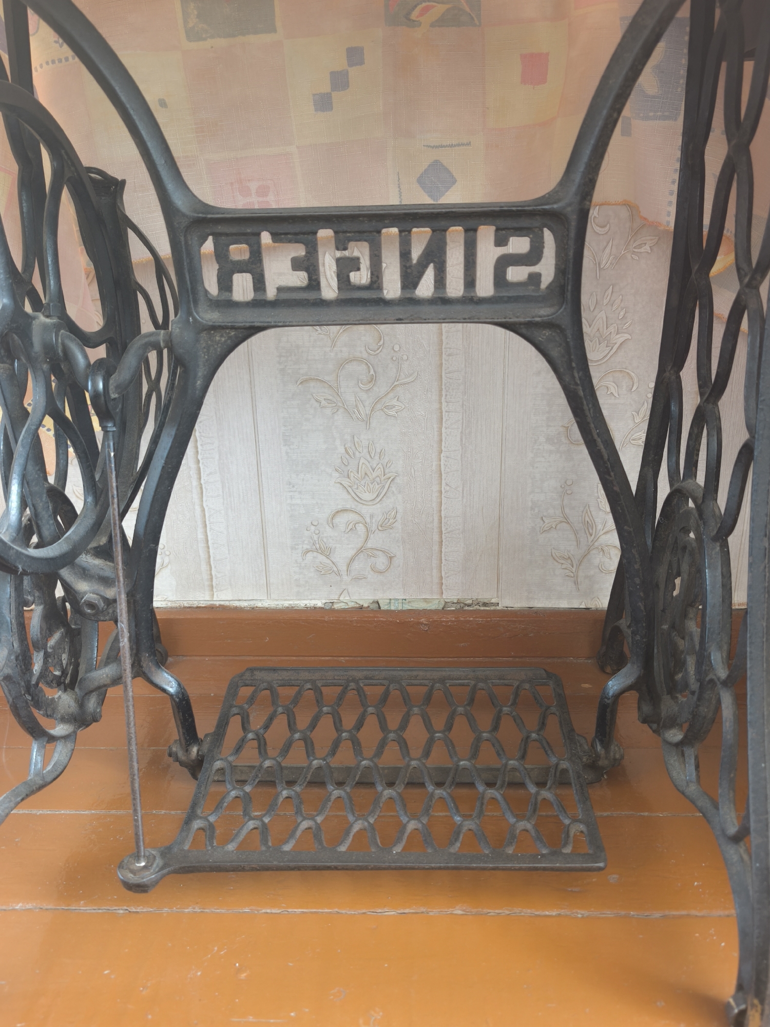 The image shows the base of an antique Singer sewing machine, likely located in Kady, Russia. The ornate cast iron frame, decorated with intricate patterns, supports a pedal mechanism. The machine stands on a polished wooden floor, and a patterned wallpaper is visible in the background. The lighting suggests an indoor setting, possibly a home or a workshop. There are no people present in the image.