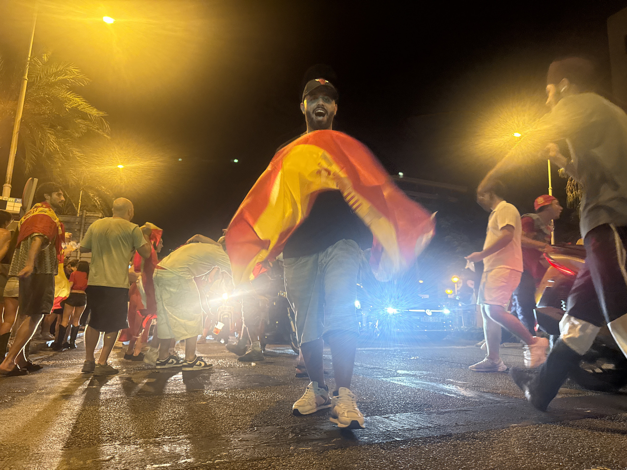 The image depicts a nighttime street scene in Barcelona, Spain, with a group of people gathered. The primary focus is a man in the center, wearing a black cap and a black shirt, holding a Spanish flag that is spread out and blurred by motion. He has an open-mouthed expression, suggesting excitement or shouting. Around him, other individuals are visible, some wearing casual clothing like t-shirts and shorts. The ground is wet, reflecting the bright, yellowish streetlights and the headlights of a vehicle in the background, indicating recent rain or a damp environment. Palm trees are visible on the left side of the frame. The overall atmosphere appears to be one of a public gathering or celebration, possibly related to a sporting event or national holiday, given the presence of the Spanish flag.