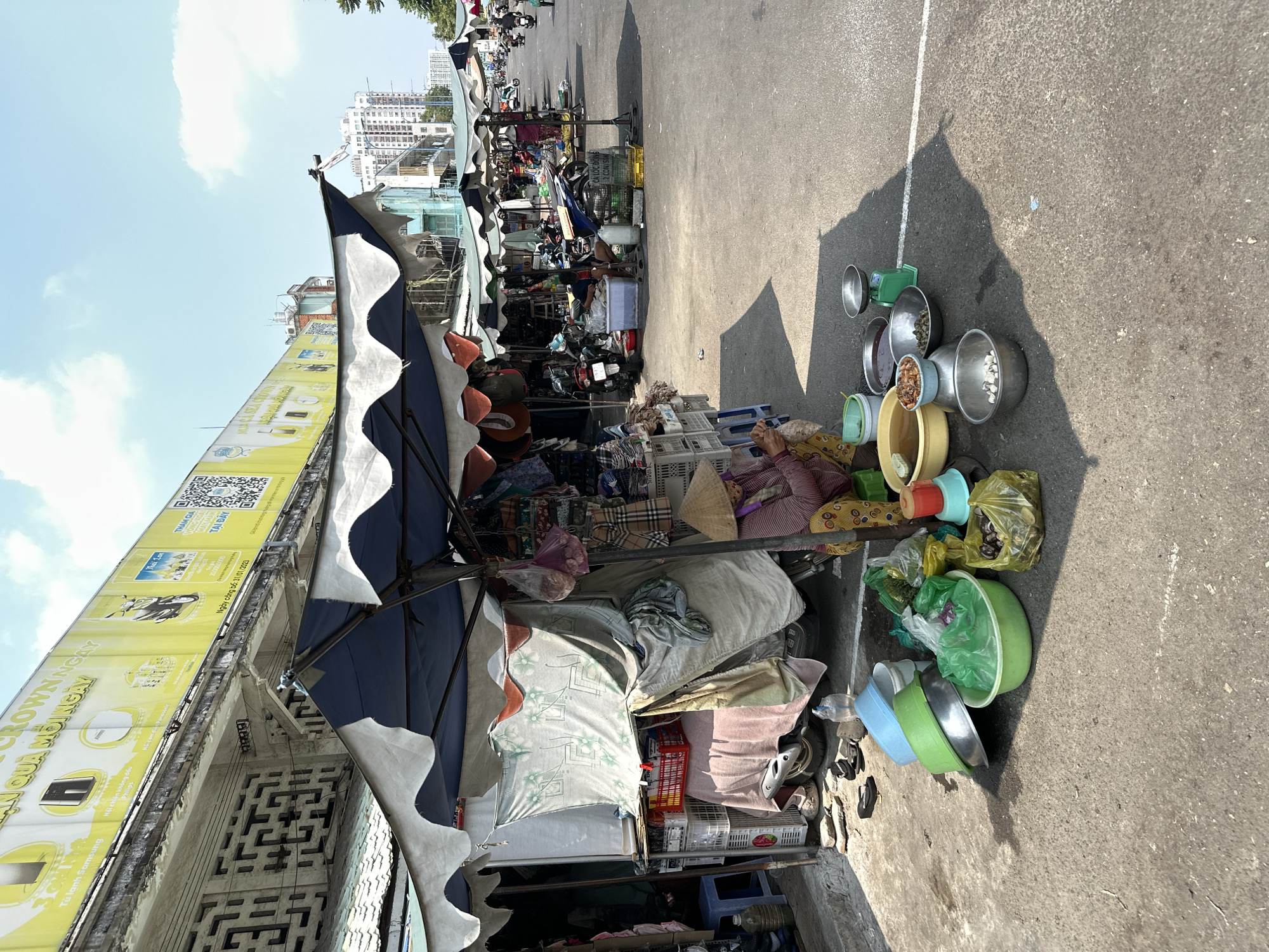 The image shows an outdoor street scene in Ho Chi Minh City, Vietnam. A makeshift stall is set up along the side of a building with a yellow billboard advertising Samsung refrigerators and other products. The stall is covered by a blue and white striped awning, with various items piled beneath, including bags, containers, and what appears to be bedding. An elderly woman wearing a conical hat is seated near the stall, surrounded by bowls of various sizes and colors, some containing food items. The street is paved, with a white line marking a boundary. In the background, other stalls and vendors are visible, along with motorcycles and buildings. The sky is partly cloudy.