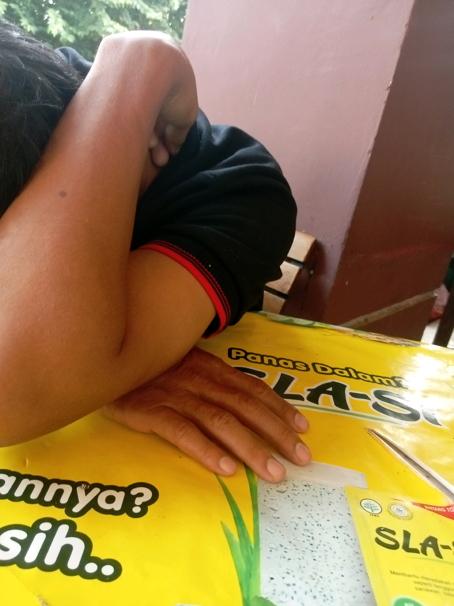 A close-up, low-angle shot shows a person's arm resting on a yellow surface, with their face obscured by their arm and hair. The person is wearing a black shirt with red trim on the sleeve. The yellow surface appears to be a packaging material with text in a dark font, including the words "Panas Dalam" and "SLA-S". The background includes a blurred outdoor scene with greenery and a pinkish-brown wall. The lighting is natural and somewhat diffused. The overall impression is of someone resting or possibly sleeping. The scene is set in Batam, Indonesia, where "Panas Dalam" (hotness within) refers to a traditional Chinese medicine concept and SLA-S is a herbal remedy brand.