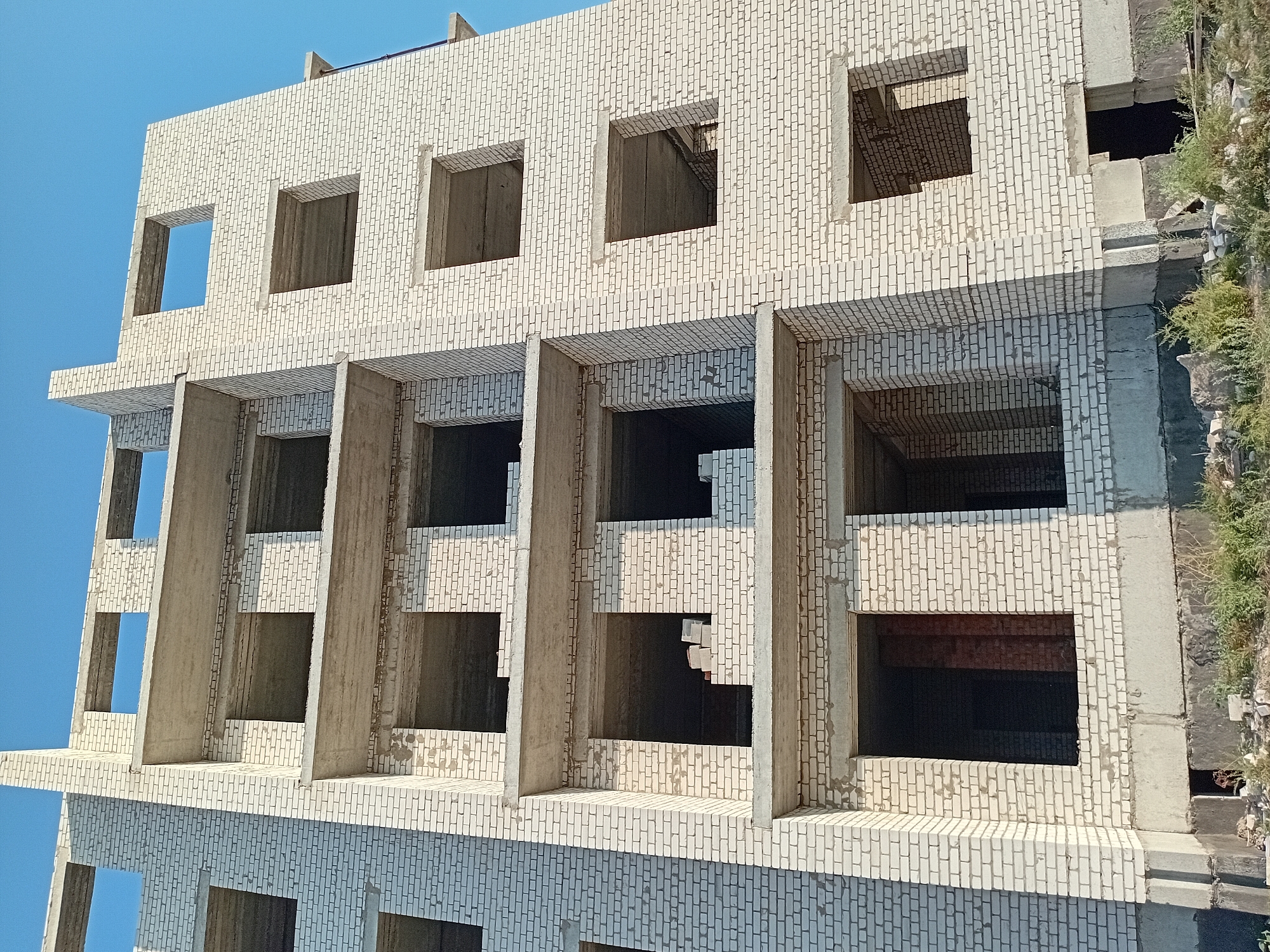 This is a forensic, matter-of-fact description of an under-construction multi-story building located in the city of Semey, Kazakhstan. The building is constructed with white brick and concrete. It features multiple levels with open, unfinished window and doorway openings. The concrete floors and ceiling slabs are visible, indicating that the structure is in an intermediate stage of construction. There is debris and vegetation at the base of the building, suggesting a construction site environment. The sky is clear and blue. No people are visible in the image.