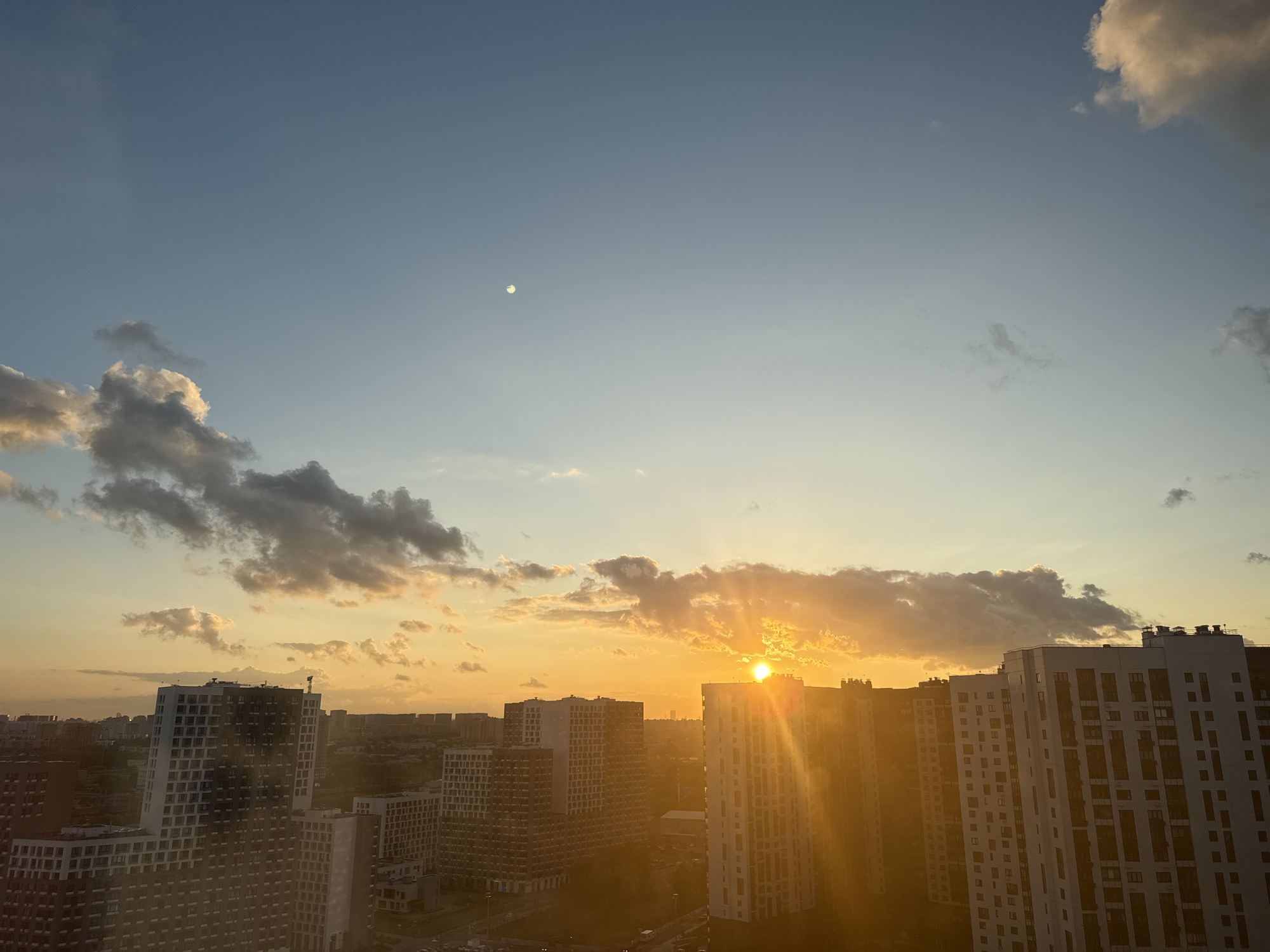 This is an eye-level, outdoor, wide shot photograph taken from a high vantage point, likely a window or balcony, overlooking a modern urban landscape during sunset. The sky is a gradient of light blue at the top, transitioning to a warm orange and yellow hue near the horizon, indicating the setting sun. The sun itself is a bright, almost blinding orb peeking through scattered clouds, casting golden rays across the scene. A faint, small, pale circle, possibly the moon, is visible in the upper left quadrant of the sky.
Dominating the foreground and midground are numerous multi-story residential buildings, characteristic of a densely populated city. The buildings are modern in design, with many featuring white and gray facades and numerous windows. Their varying heights and architectural styles create a layered effect. The sunlight glints off the surfaces of these buildings, creating bright highlights and casting long shadows. The sun's rays are so intense that they create a lens flare effect across the image, particularly on the right side.
In the distance, a sprawling cityscape is visible, suggesting the vastness of Lyubertsy, Russia. The silhouette of more buildings and structures can be seen against the vibrant sunset sky. There are no visible people or vehicles in the frame, suggesting the photograph was taken at a time when activity was minimal or the viewpoint was too distant to discern them. The environment appears to be a typical residential district within a large Russian city. The overall impression is one of urban development bathed in the dramatic light of a sunset.