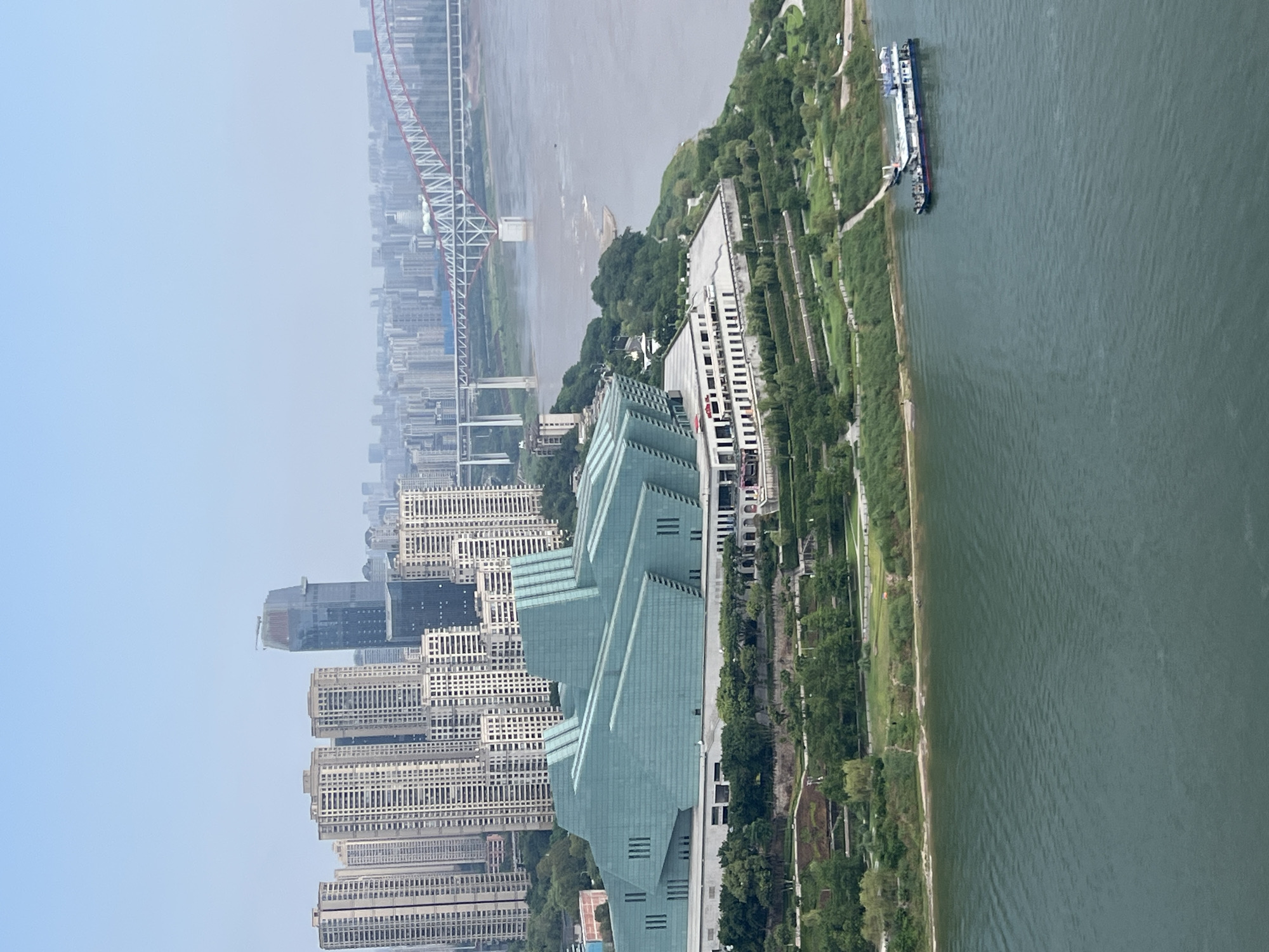 This aerial photograph, captured in Yuzhong District, China, showcases a vibrant cityscape situated along a wide, calm river. Dominating the foreground is a striking, multi-tiered modern building with a distinctive angular, teal-colored glass facade, nestled on a verdant, terraced hillside. The building's architecture is characterized by its geometric shapes and large glass panels, reflecting the overcast sky.
Beyond the modern structure, a dense collection of tall, light-colored residential buildings forms the urban backdrop, stretching towards the horizon. In the distance, a massive, arching bridge with red and white detailing spans the river, connecting different parts of the city. The river itself, a murky green, occupies the lower portion of the frame. A long, multi-decked vessel with a blue and white superstructure is seen navigating the water near the bank, just in front of the terraced landscape. The sky is a pale, hazy blue, suggesting a slightly diffused light. The overall impression is one of a bustling urban center integrated with its natural riverine environment.