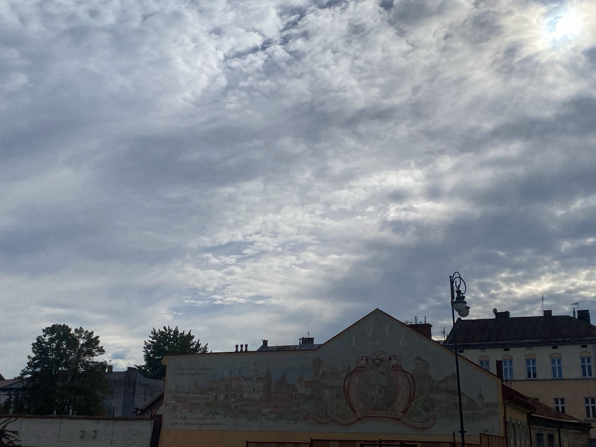 The image shows a cloudy sky over buildings in the city of Nowy Sącz, Poland. In the foreground, there is a mural on the side of a building. The mural depicts a historical scene with buildings, trees, and a crest. The year "1946" is visible above the crest. A street lamp with ornate detailing stands in front of the building with the mural. The background shows more buildings and trees. The overall atmosphere is somewhat overcast, with the sun attempting to break through the clouds. The image is taken from a low angle, looking up towards the sky and the tops of the buildings.