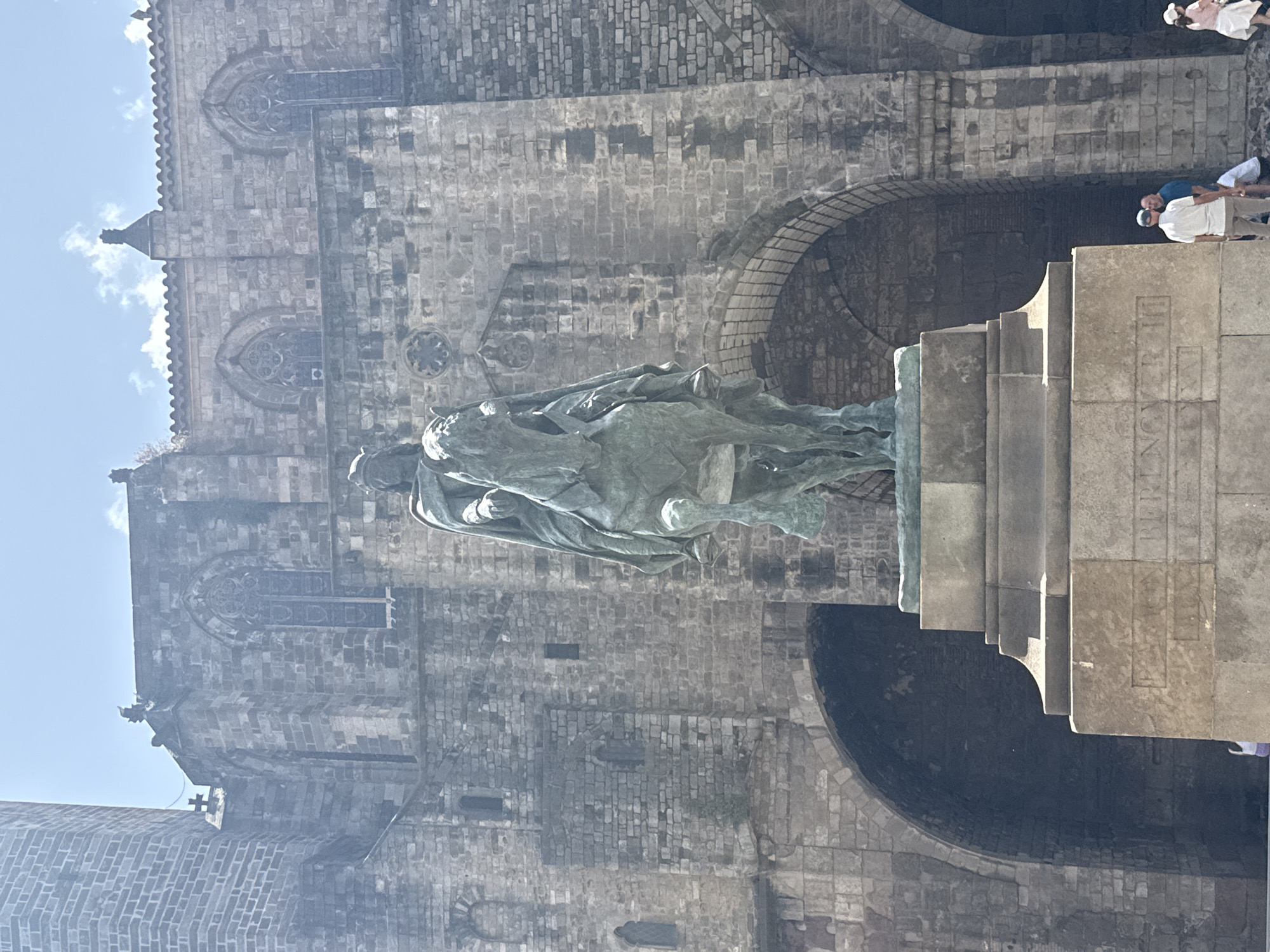 The image shows a full-body, eye-level shot of a bronze equestrian statue in front of a stone building. The statue depicts a person in armor and a cape riding a horse. The building behind the statue has arched doorways and windows with intricate stonework. The sky above is blue with a few clouds. In the foreground, the base of the statue has inscriptions. To the right of the statue, a few people are standing and walking. The statue is located in Barcelona, Spain.