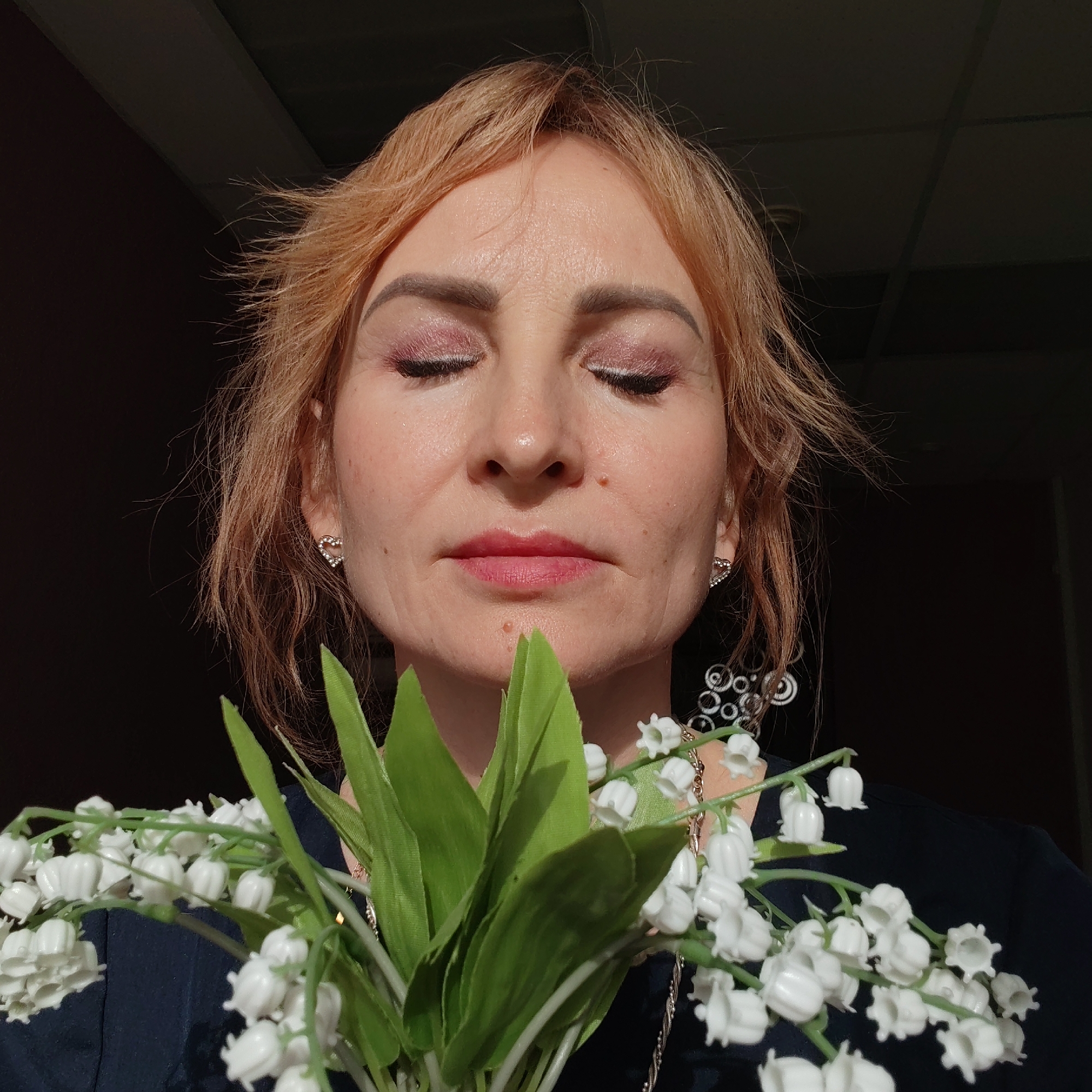A middle-aged woman is shown in a close-up shot holding a bouquet of lily of the valley flowers with green leaves. The woman has fair skin, light reddish-blonde hair, and is wearing makeup, including pink eyeshadow. Her eyes are closed, and she has a serene expression. She is wearing heart-shaped earrings.
She is in what appears to be an indoor setting, with a dark background behind her. The woman wears a dark colored shirt. It is impossible to determine the exact location in Chaikovsky, Russia, from the given image.