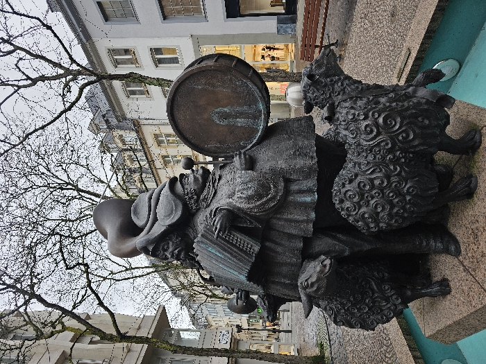 A bronze sculpture depicting a group of stylized musicians and sheep is positioned centrally on a stone base featuring a teal-colored water basin. The sculpture consists of three primary figures: a prominent individual playing an accordion, another holding drumsticks poised over a large drum, and a third figure wearing a hat with a horn-like protrusion. Four detailed sheep with textured, curly wool are depicted standing around the base of the human figures.
The scene is located on Place d'Armes, Luxembourg City, Luxembourg. In the background, multi-story buildings with light-colored facades and traditional European architectural elements line a paved street. Deciduous trees with bare branches stand against an overcast, grey sky, suggesting a cold season. The textured pavement appears damp, indicative of recent precipitation. A brown wooden slatted park bench is visible to the right of the sculpture. No active human presence is discernible in the immediate foreground or midground.