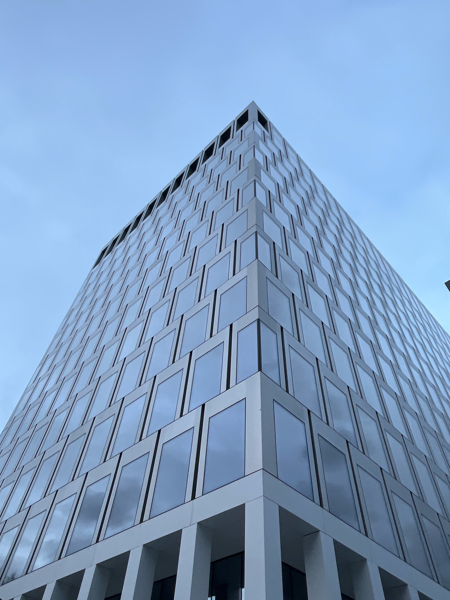 The image shows a modern, sharp-angled skyscraper in Moscow, Russia, viewed from a low angle. The building has a facade composed of many rectangular windows, framed by white architectural elements. The sky above is a muted blue with some cloud cover. The building's structure creates a sense of immense scale and geometric precision. There are no people or vehicles visible in the frame. The overall impression is one of urban architecture and design.