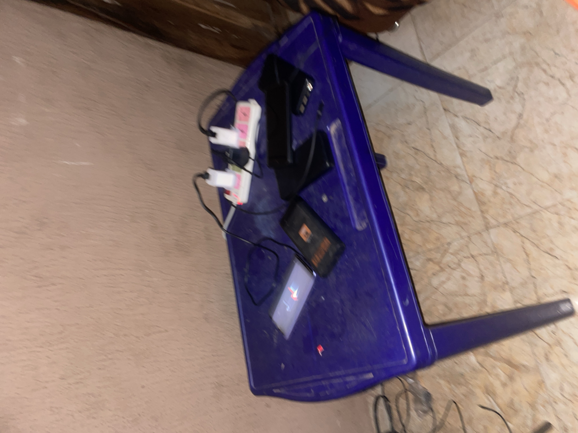 On a purple plastic table in Lokoja, Nigeria, a charging strip with two white power adapters and pink electrical outlets is connected to two phones. A black phone with a red charging icon is on the left, and a second black phone is on the right. A charging cable is also present. The table is situated on a marbled floor, with a textured, light brown wall in the background.