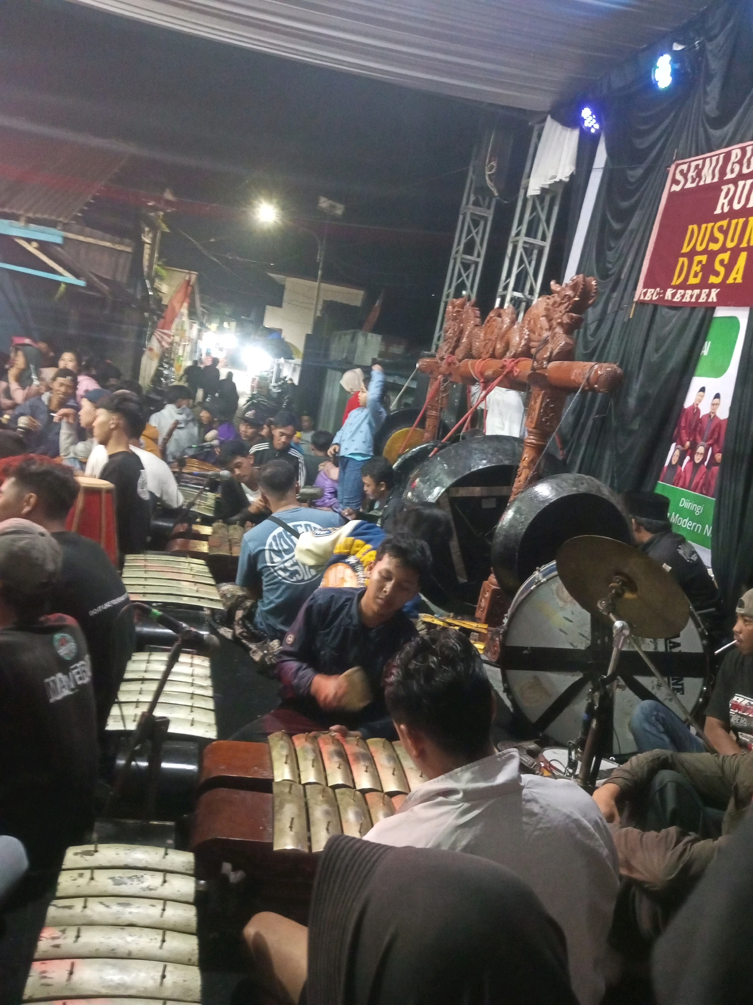 The image depicts an outdoor gathering with a gamelan orchestra performing at night in Wonokromo, Indonesia. The scene is illuminated by artificial lights, with a large black tarp serving as a backdrop for the performance. A banner with text in Indonesian, including "SENI BUDAYA RUMANI DUSUN DE SA KEC: KERTEK," indicates the event's cultural nature. Several individuals are present, some actively engaged in playing the gamelan instruments, while others appear to be spectators. The instruments include large gongs, a drum, and a keyed metallophone. The environment suggests a festive or traditional community event.