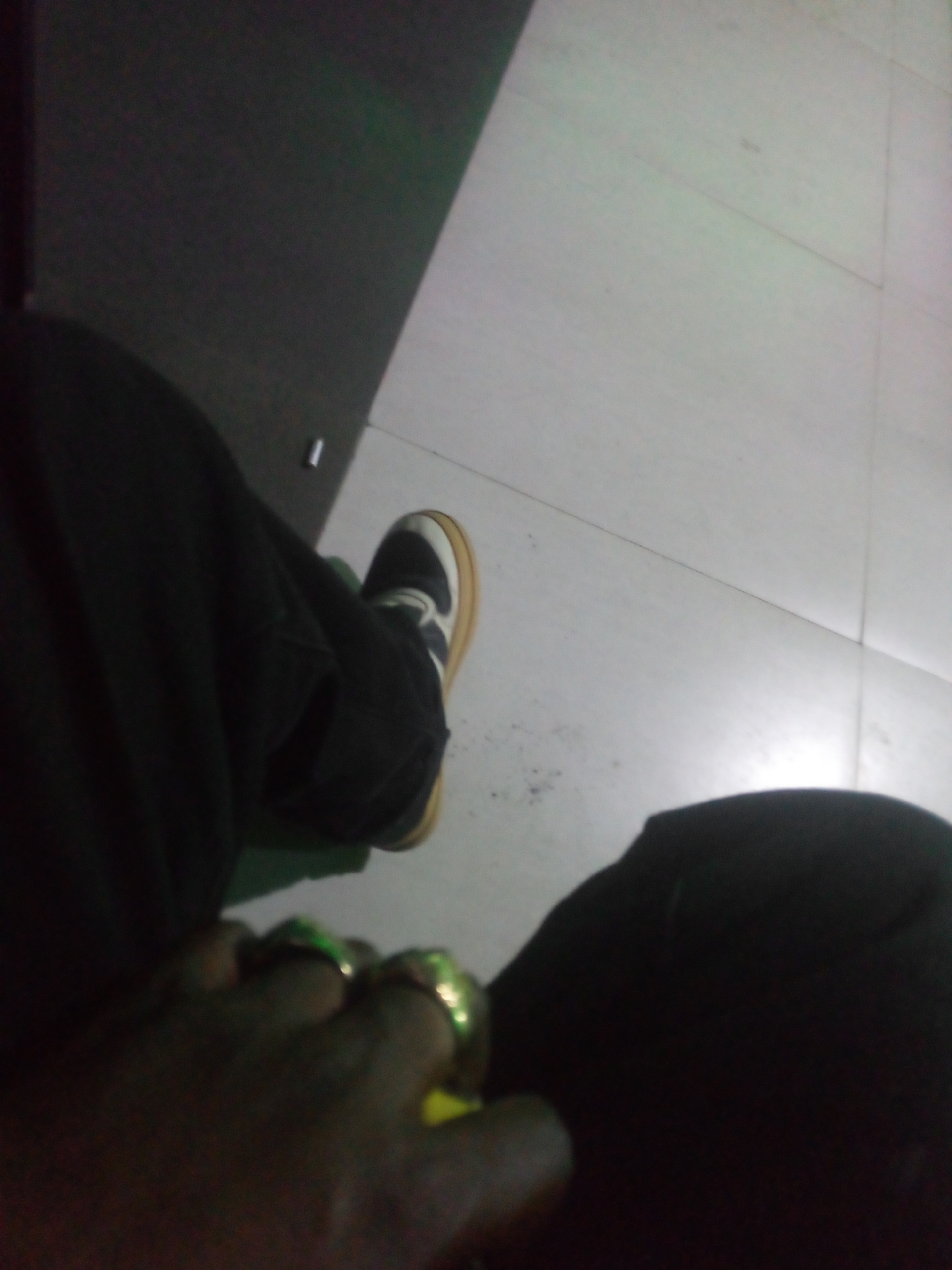 The image is a first-person perspective of a person sitting, with their legs and feet visible. The person is wearing dark trousers and a black and white sneaker with a tan sole. The person is also wearing rings on their fingers, which appear to be gold with green stones. The environment is indoors, with a tiled floor that is light in color and a darker, textured wall or surface in the background. The lighting is dim. The image appears to have been taken in Jos, Nigeria.