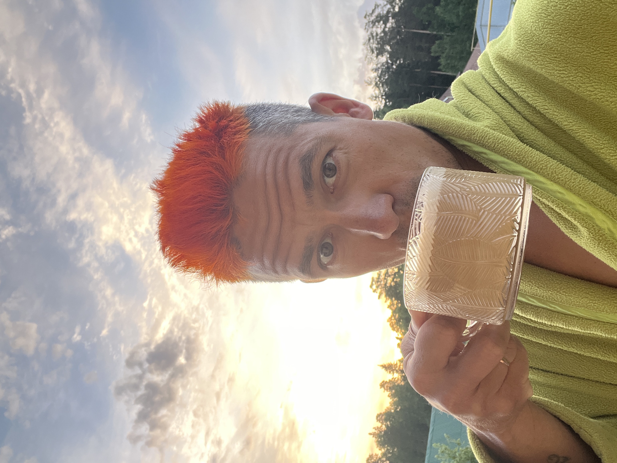 A man with short, dyed-orange hair is holding a glass of a beverage in front of a backdrop featuring a cloudy sky, trees, and buildings. The man is wearing a lime green robe. He is looking directly at the camera. He is holding the glass with his right hand. The ring finger of his right hand has a ring on it. It is daytime. The photo was likely taken in Chaikovsky, Russia.