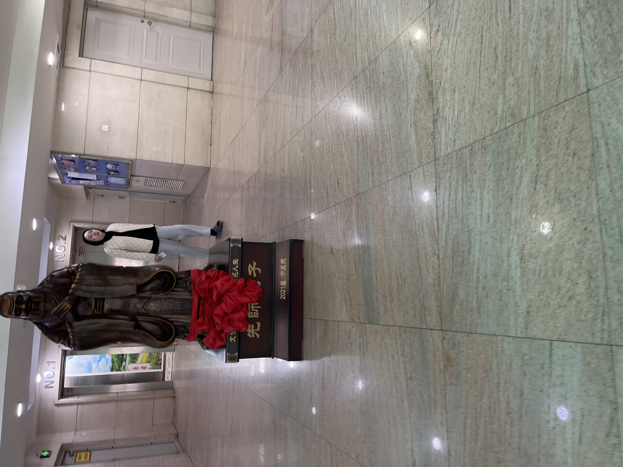 The image captures the interior of a building in Changchun City, China. A woman stands adjacent to a statue of a sage. The statue is adorned with a red rosette and sits atop a plinth inscribed with Chinese characters: 先師子 (First Teacher Zi), 2021届于昊天 (2021 Class Yu Haotian), and 文光人生(Bright life). The background includes a door, a notice board, and elevators. The floor is tiled.