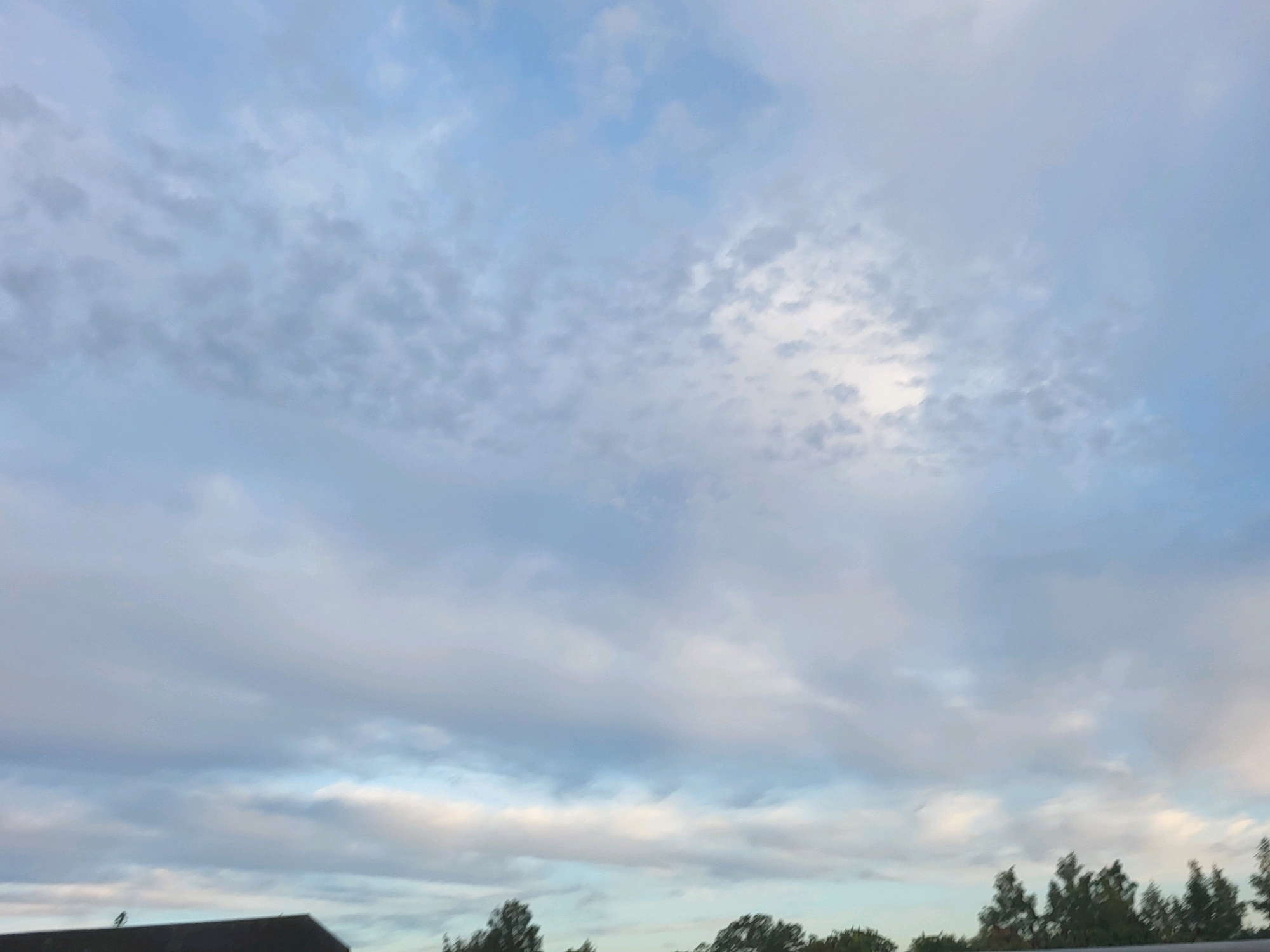 The image shows a cloudy sky with a few trees and rooftops visible at the bottom. The sky is predominantly light blue with varying shades of white and gray clouds. Some areas of the sky are brighter, suggesting sunlight filtering through the clouds. The clouds are spread across the frame, with some appearing thinner and wispy, while others are more dense and layered. A dark roof of a building is partially visible in the lower left corner, and a line of trees with dark green foliage runs along the bottom edge of the image. The scene appears to be an outdoor view from ground level looking upwards towards the sky.