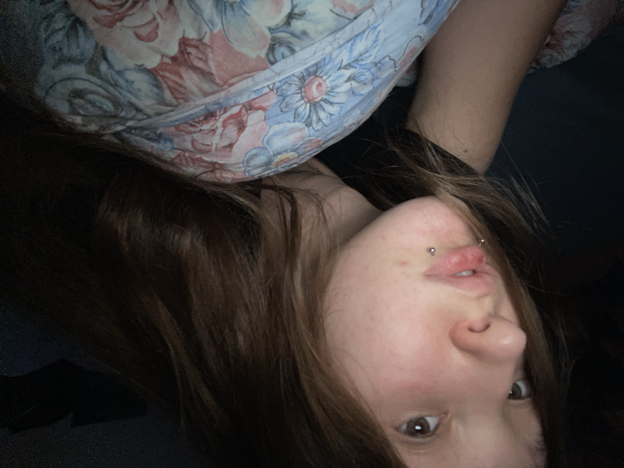 A young woman with light brown hair is lying down, partially covered by a floral-patterned blanket. She is looking off to the left with a neutral expression. She has a lip piercing. The background is dark and appears to be indoors.