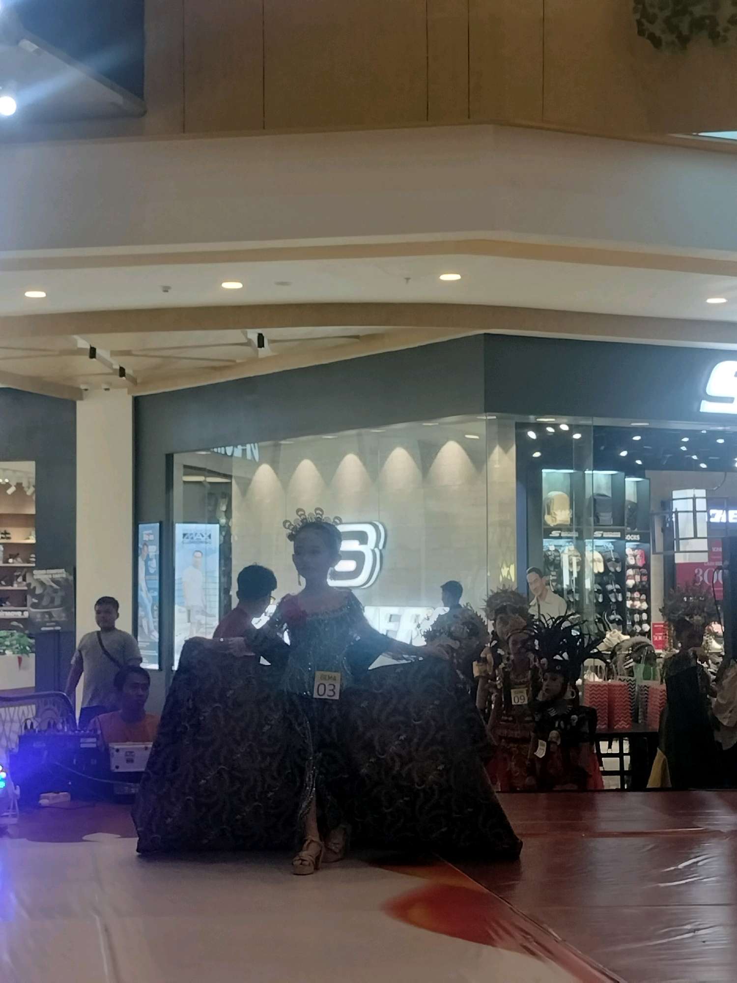The image depicts a fashion show event taking place indoors, likely within a shopping mall in Batam, Indonesia. The primary focus is on a young woman in an elaborate traditional costume, possibly a kebayak, with ornate headwear, standing on a stage. She is wearing heels and has a numbered tag "03" on her dress.
Several other individuals are visible in the background. To the left, a man wearing a grey t-shirt and a backpack stands near a sound system with blue lights. Another person, wearing an orange t-shirt, is seated behind the sound equipment. Further back, there are other participants in similar festive attire, with elaborate headpieces and costumes. Behind them, storefronts with glass facades are visible, one displaying a large "B" logo and another featuring various products and mannequins. The overall setting suggests a public event or competition within a commercial space. The lighting appears to be artificial, typical of indoor environments.