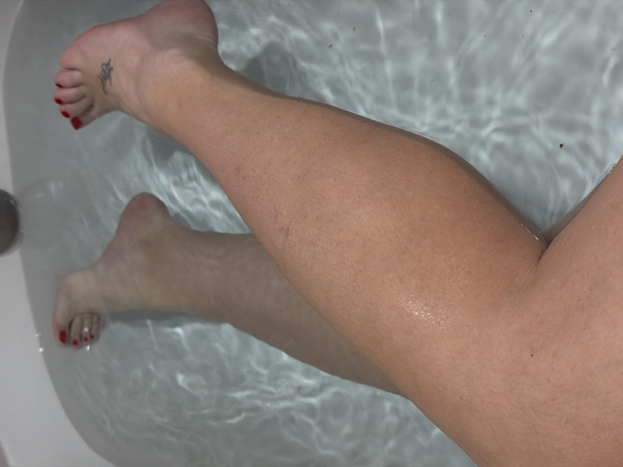 A photograph taken from above depicts a pair of legs submerged in water within a white bathtub. The feet have red nail polish and one foot has a small tattoo. The water is clear and the legs appear fair-skinned with some visible hair follicles. The environment is a bathroom. The location of the photograph cannot be determined to be in Chaikovsky, Russia, based solely on the image content.