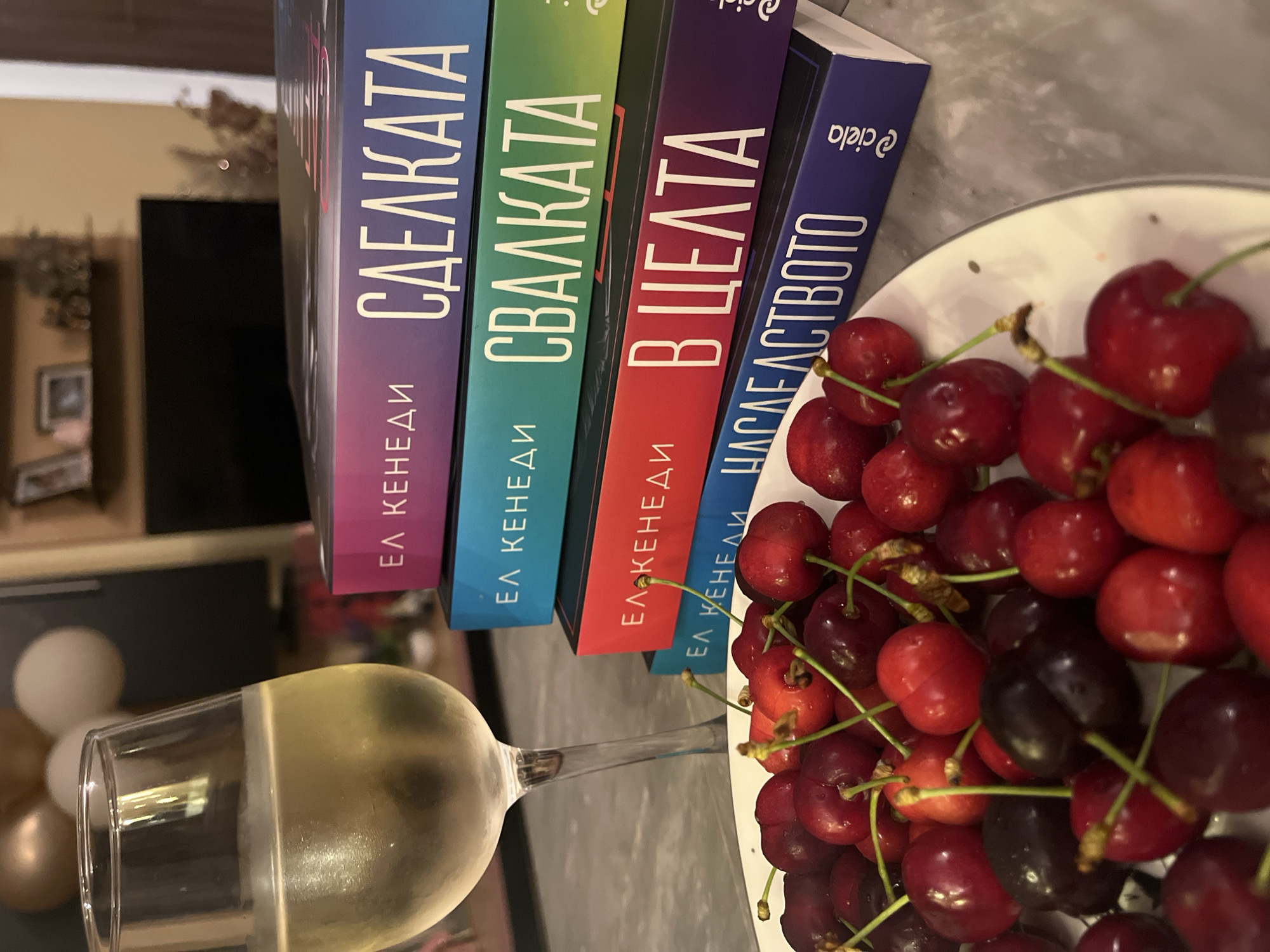 This indoor still life photograph captures a relaxing moment, presented with a 90-degree counter-clockwise rotation, revealing books, fresh fruit, and a beverage. The scene is located in Veliko Tarnovo, Bulgaria.
On the left side, a clear wine glass filled with a light, yellowish liquid, likely white wine or champagne, is prominently featured. To its right, a stack of five books is arranged, showcasing their colorful spines. From top to bottom in the rotated image (or left to right if viewed upright), the books are:
1. **Purple spine:** "ЕЛ КЕНЕДИ СДЕЛКАТА" (Elle Kennedy, The Deal)
2. **Teal spine:** "ЕЛ КЕНЕДИ СВАЛКАТА" (Elle Kennedy, The Mistake)
3. **Green spine:** Displays "ЕЛ КЕНЕДИ" (Elle Kennedy), with the title partially obscured.
4. **Red spine:** "ЕЛ КЕНЕДИ В ЦЕЛТА" (Elle Kennedy, The Goal)
5. **Dark blue spine:** "ЕЛ КЕНЕДИ НАСЛЕДСТВОТО" (Elle Kennedy, The Legacy), also featuring the publisher's logo "@ciela".
Next to the books, a white plate is abundantly filled with ripe cherries. The majority are a vibrant red, interspersed with darker, almost black cherries, many still bearing their slender green stems.
The background is softly blurred, indicating a shallow depth of field. Visible elements include a dark, rectangular object (possibly a television screen) and hints of light-colored, spherical objects, perhaps white and gold balloons, suggesting a cozy living space. The ambient indoor lighting contributes to a calm and inviting atmosphere.