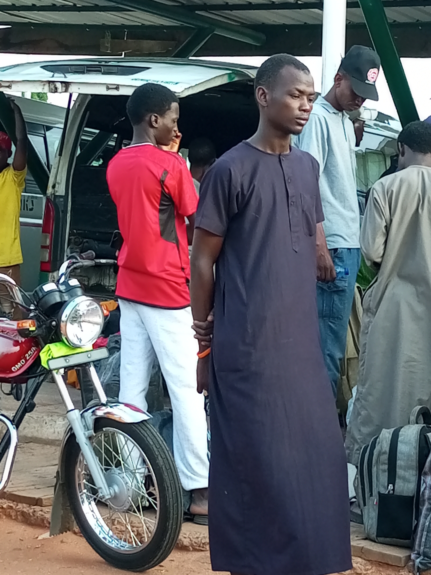 In Gombe, Nigeria, a group of men stand near a van and a motorcycle under a covered structure. The man in the foreground wears a dark navy, ankle-length garment with a quarter-length placket and buttons. To his left, a man in a bright red t-shirt with dark accents on the sleeves and sides stands next to a white van, his back partially turned. He wears light grey pants. Behind him, a man in a light grey t-shirt and blue jeans, wearing a black baseball cap, is also visible. To the far right, a man in a light tan, long-sleeved garment stands with a gray backpack at his feet. A red motorcycle is parked in the foreground on the left, with its front wheel and fender clearly visible. The motorcycle has "GMO 25IN" printed on its side. The ground is reddish-brown dirt, and the structure above is supported by metal poles and has a corrugated metal roof. The scene appears to be an outdoor waiting area or transportation hub.