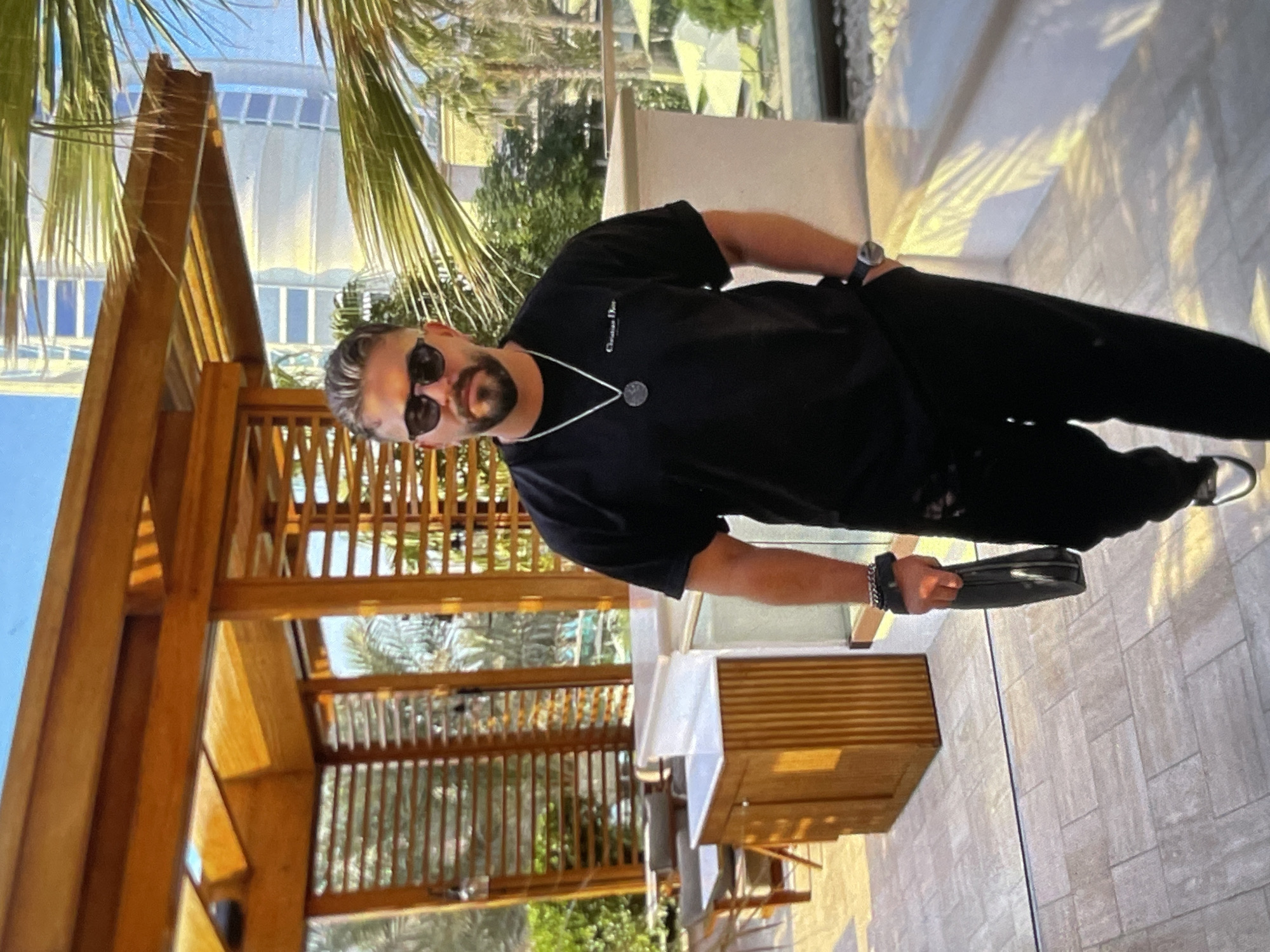 A medium shot captures a man walking outdoors in what appears to be a resort or upscale outdoor dining area. He is positioned in the center of the frame, facing slightly to the right. The man is wearing a black t-shirt with a small white logo that reads "Christian Dior," black pants, and black sunglasses. He has dark hair and a beard, and is wearing a silver chain with a circular pendant. On his left wrist, he sports a watch and a thick black bracelet. He is holding a black clutch bag in his left hand.
The background suggests a sunny day. To the left, there's a wooden structure with a slatted roof and vertical wooden slats, possibly a pergola or covered seating area, with tables and chairs visible. Beyond that, palm trees and other greenery are present. To the right, a modern building with numerous windows can be seen, along with more palm trees and what look like outdoor umbrellas. The ground is paved with light-colored tiles. The lighting indicates it is daytime, with sunlight casting shadows from the wooden structure and palm fronds onto the ground and walkway.