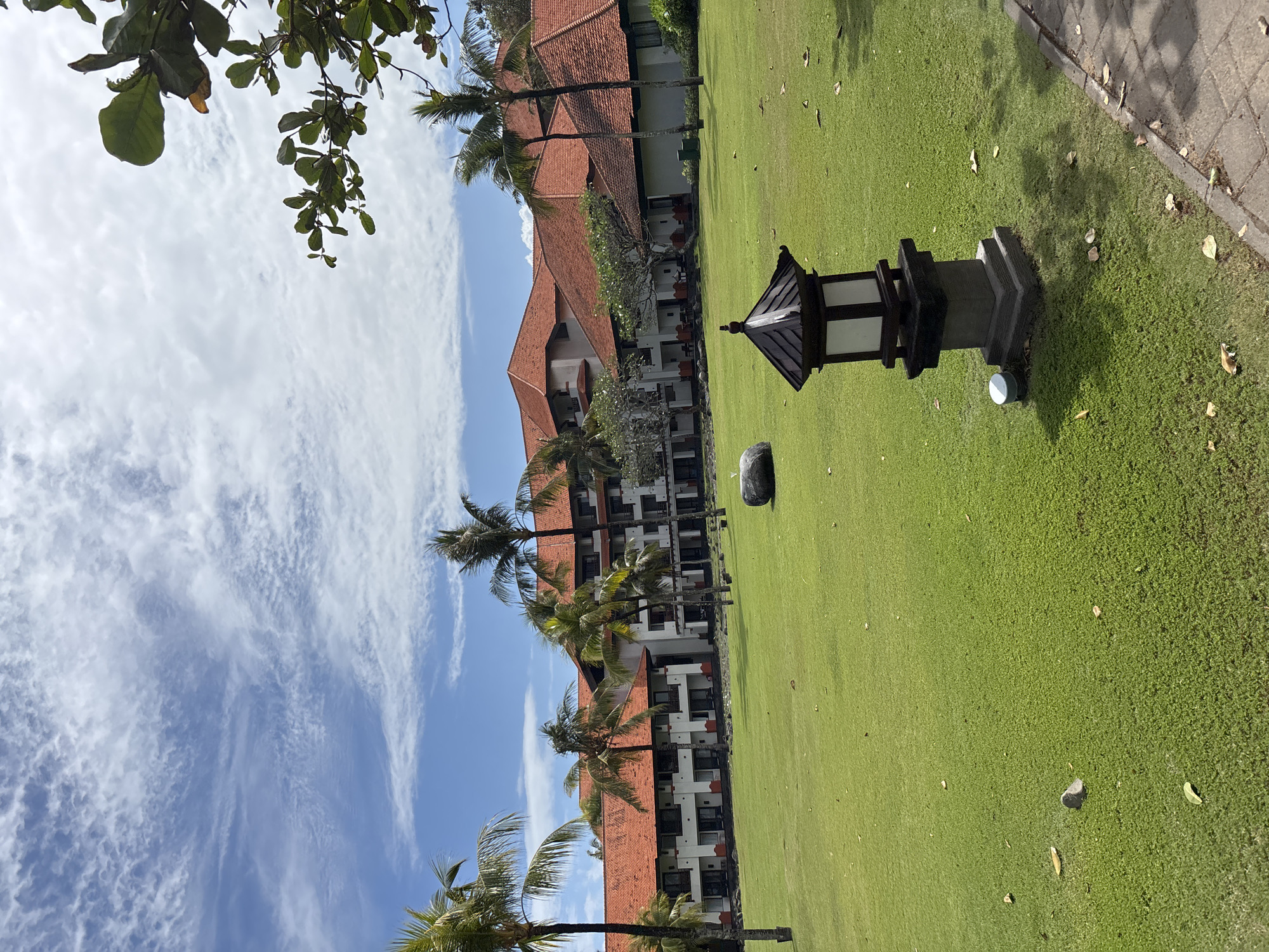 The image depicts a resort property in Kuta Selatan, Indonesia, on a partly cloudy day. In the foreground, a paved walkway runs along the right side of the frame, bordering a neatly manicured, vibrant green lawn. A decorative, dark-colored lantern with a tiered base stands on the lawn near the walkway. Scattered across the lawn are a few fallen leaves and a small, greyish rock.
In the midground, a large, multi-story resort building with distinctive red-tiled roofs stretches across the frame. The building features white walls, multiple balconies, and dark-trimmed windows. Several palm trees, with their characteristic fronds, are interspersed around the building and on the lawn, suggesting a tropical setting. One larger, dark rock is visible on the lawn between the resort and the lantern.
The background is dominated by a bright blue sky with scattered white, wispy clouds. The upper right portion of the image shows the green leaves and branches of a tree extending into the frame from above. No people are visible in the scene.