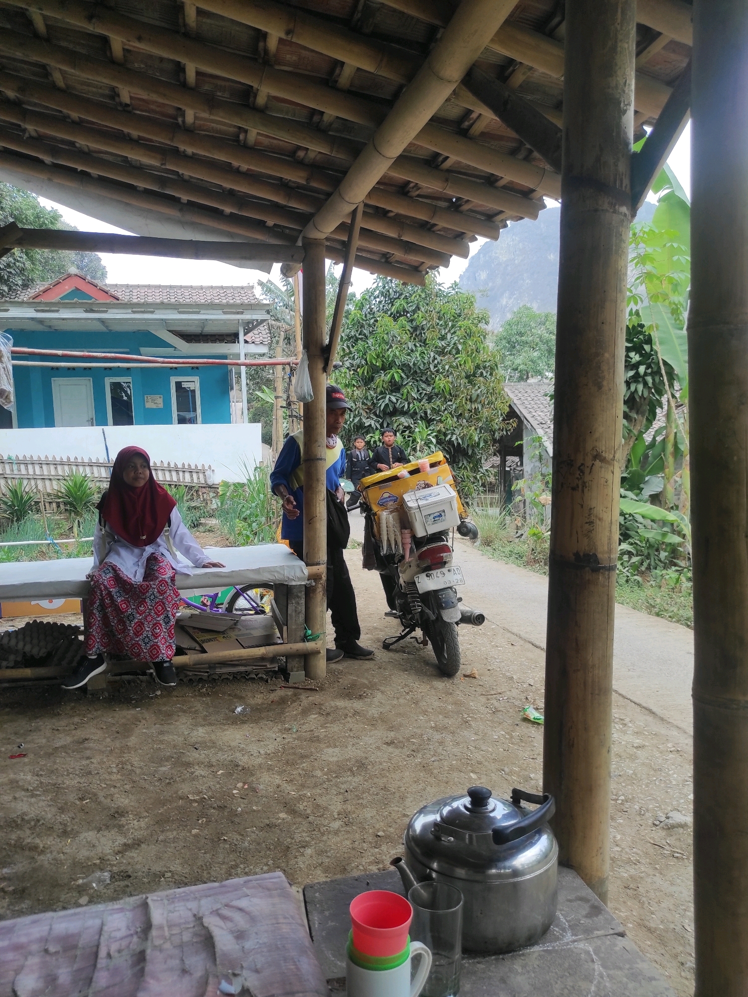 The image depicts an outdoor scene under a bamboo-covered shelter, likely in Gunungmasigit, Indonesia. In the foreground, a young girl wearing a maroon hijab and a patterned skirt sits on a long, white-covered surface, possibly a table or bench. To her right, a man in a blue shirt and dark pants stands near a motorcycle. The motorcycle is yellow and has a large storage box mounted on the back, filled with white coolers and stacks of cups. Behind the people and motorcycle, a bright blue house with white trim is visible, along with a white fence and lush green vegetation. In the distance, a mountain can be seen. The ground is dirt and gravel, and the shelter's roof is constructed from bamboo poles and slats. On a table in the lower right corner, a silver teapot and several cups are present. The overall impression is a casual, roadside setting.