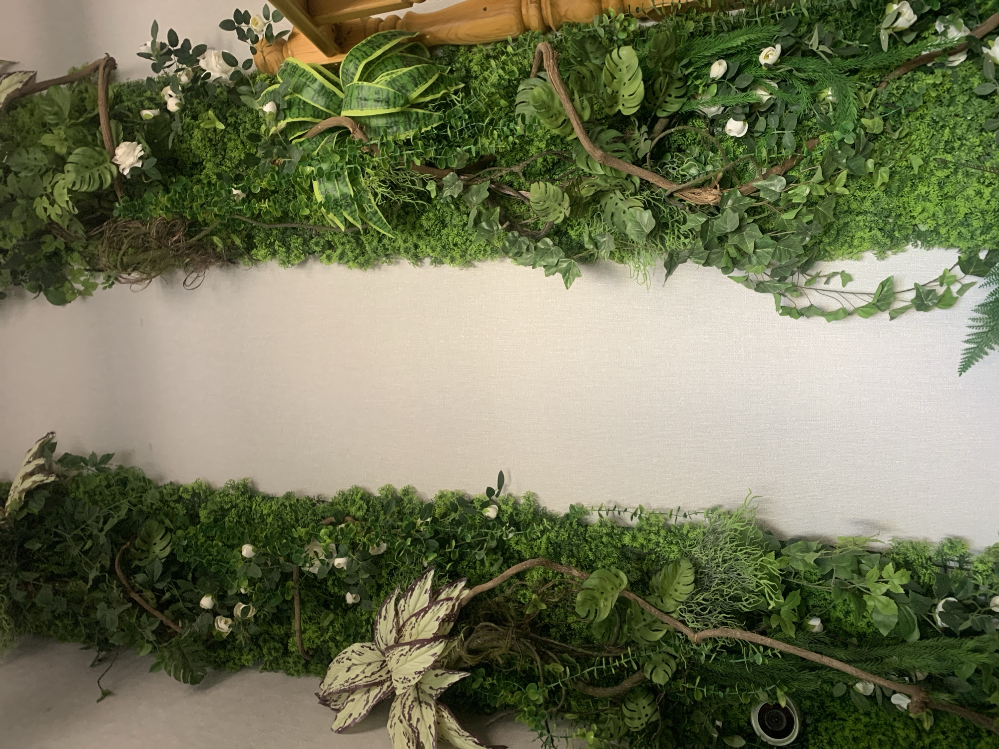 The image shows a vertical garden installation on a white wall. The garden consists of various artificial green plants, including moss, ferns, ivy, and some flowering plants with small white blossoms. Two prominent vertical sections of dense greenery are positioned on either side of the frame, with a lighter wall space in between. On the left side, a large, light-colored leaf with dark markings is visible. On the right side, a wooden railing of a staircase is partially visible at the edge of the frame. The overall impression is one of an indoor decorative element designed to mimic a lush, natural environment. There are no people or any specific architectural details that would allow for identification of a location within the city of Almaty, Kazakhstan.