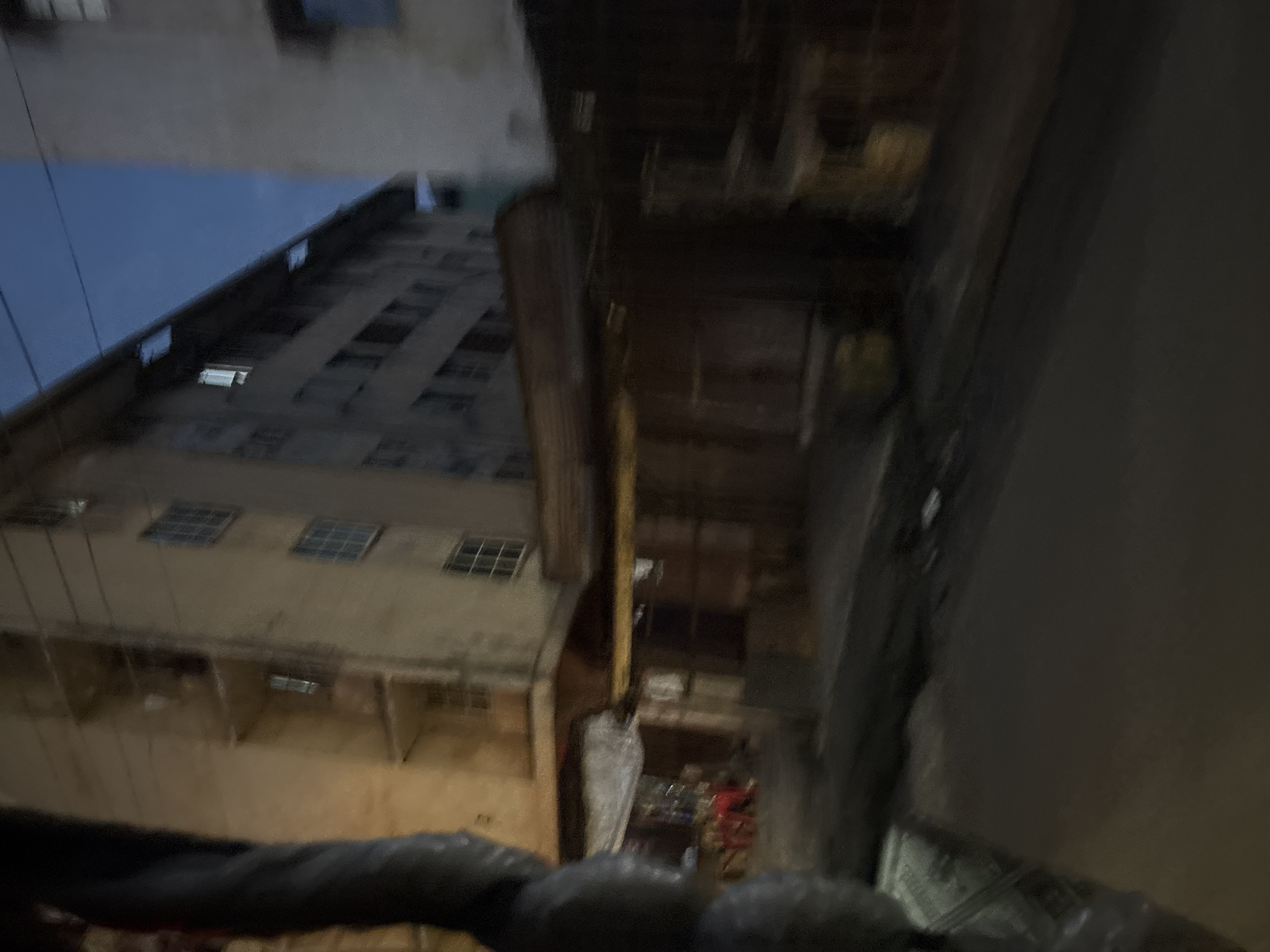 The image depicts a street scene at dusk in Onitsha, Nigeria, with a multi-story building dominating the background. The building appears to be an apartment complex or office building, with multiple windows visible on its facade. In the foreground, there are what appear to be market stalls or shops lining the street, with awnings providing shelter. Some individuals can be seen near the stalls, though their features are indistinct due to the low light and motion blur. The street surface is asphalt, and there are signs of pedestrian activity. The overall atmosphere is that of a busy urban environment at the end of the day, with the transition from daylight to darkness.