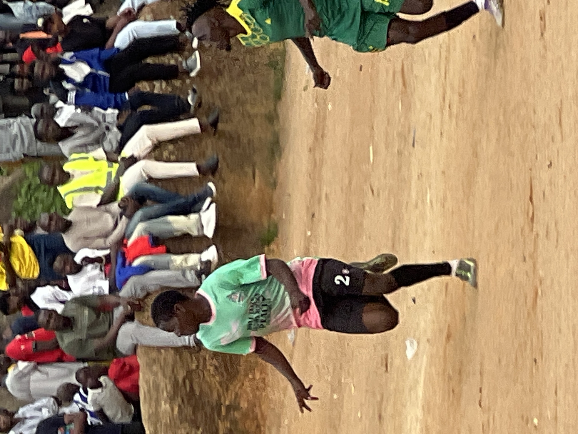 In an outdoor scene on a dirt field, likely in Jos, Nigeria, two young men in soccer attire are the primary subjects. The man on the left, wearing a green and pink jersey with the number "2" visible on his black shorts, is in mid-stride, his body angled towards the right. His left arm is extended, and his right arm is bent at the elbow. The man on the right, also in soccer attire, is in motion, his right arm extended and his head turned to his left, looking away from the camera. Behind them, a group of spectators are seated on a raised dirt embankment. They are dressed in casual clothing, with some wearing face masks. The environment is dry and dusty, with sparse vegetation in the background. The lighting suggests it is daytime, with soft shadows. The overall atmosphere appears to be one of a casual sporting event.