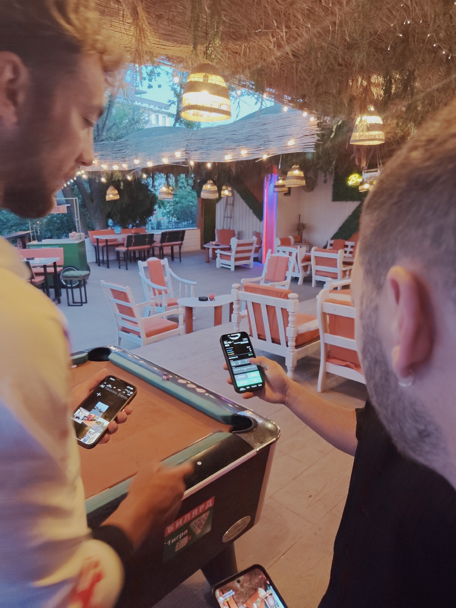 In Tsarevo, Bulgaria, at an outdoor venue with a thatched roof and string lights, two men are visible. One man, in the foreground on the left, wearing a white shirt, holds a smartphone displaying photos. He stands next to a billiard table. The second man, in the foreground on the right, wearing a black t-shirt, holds a smartphone showing a black interface with text and green buttons. The background reveals an outdoor seating area with tables and chairs, some with orange cushions, and foliage. A neon pink light is visible in the distance. The overall scene suggests a casual, social setting.