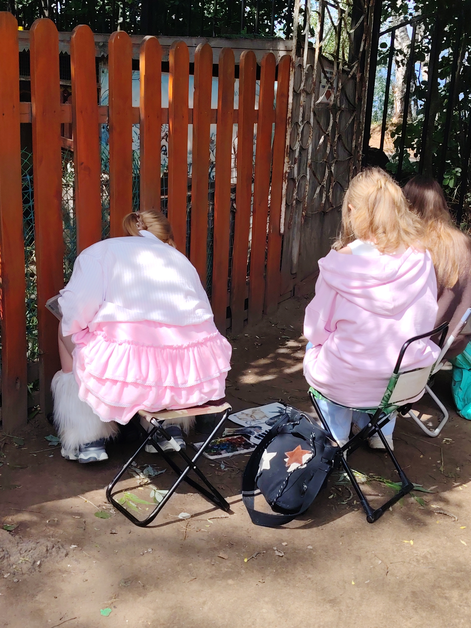 Three individuals, seen from behind, are seated outdoors in Chaikovsky, Russia, on what appears to be a sunny day with clear skies. They are positioned along a vibrant orange wooden fence and a weathered metal gate with ornate patterns. The ground is dirt, with scattered leaves.
The person on the left wears a white top over a distinct, layered pink ruffled skirt and white furry boots. They are seated on a small, dark-framed portable stool, seemingly engaged in an activity like drawing or painting, as hinted by paper or a board near their lap. The person in the middle wears a light pink hooded sweatshirt and light-colored pants, seated on a portable chair with a patterned fabric seat. A black bag with a white and orange star design lies on the ground next to this chair, along with what appears to be a paint palette featuring various colors and some papers or canvases. The third person on the right is partially visible, wearing a brown top, also seated on a similar portable chair, further indicating a group activity.
The setting suggests an art session or a leisurely outdoor gathering in a natural environment, with lush green foliage visible behind the fence and gate. Shadows on the ground suggest it is likely midday or afternoon. No legible text is visible in the image. The overall impression is one of calm, focused activity in a pleasant outdoor space.