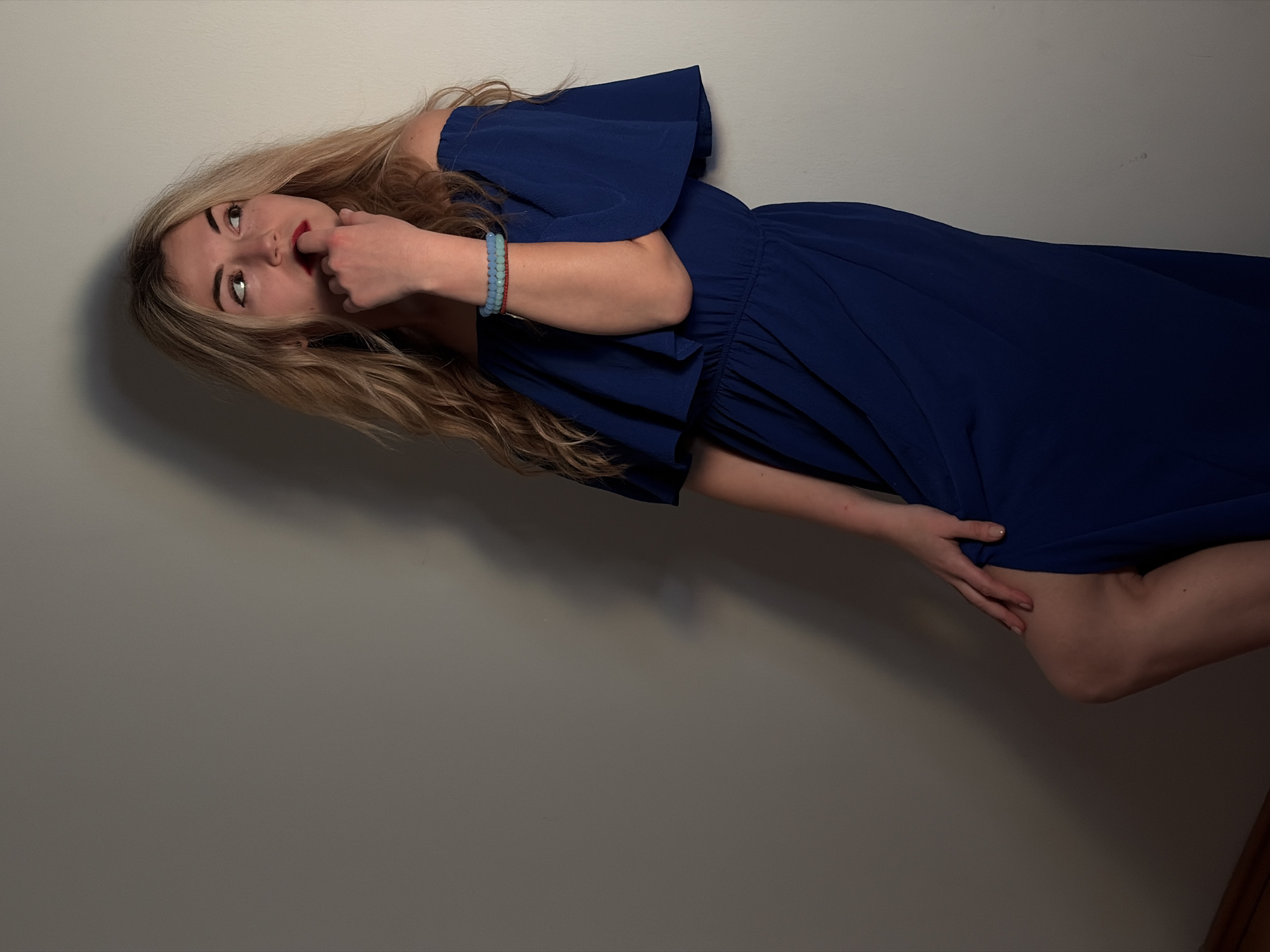 A single adult female is depicted standing indoors, leaning against a plain, light-colored wall. She has long, wavy blonde hair and is wearing a royal blue off-the-shoulder maxi dress with an elasticized waist and a high slit revealing her left knee and upper thigh. Her right hand is positioned with fingers (index finger and thumb) touching her lower lip, while her left hand rests on her bent left knee. On her right wrist, two beaded bracelets are visible: one light blue/turquoise and one red. She wears red lipstick and her gaze is directed upwards and to the right. A dark shadow is cast on the wall behind her, primarily to her left side and above her head, indicating a light source from the viewer's front-left. A faint wooden baseboard is visible at the bottom left. The image contains no discernible features to indicate a specific location within Budva, Montenegro.