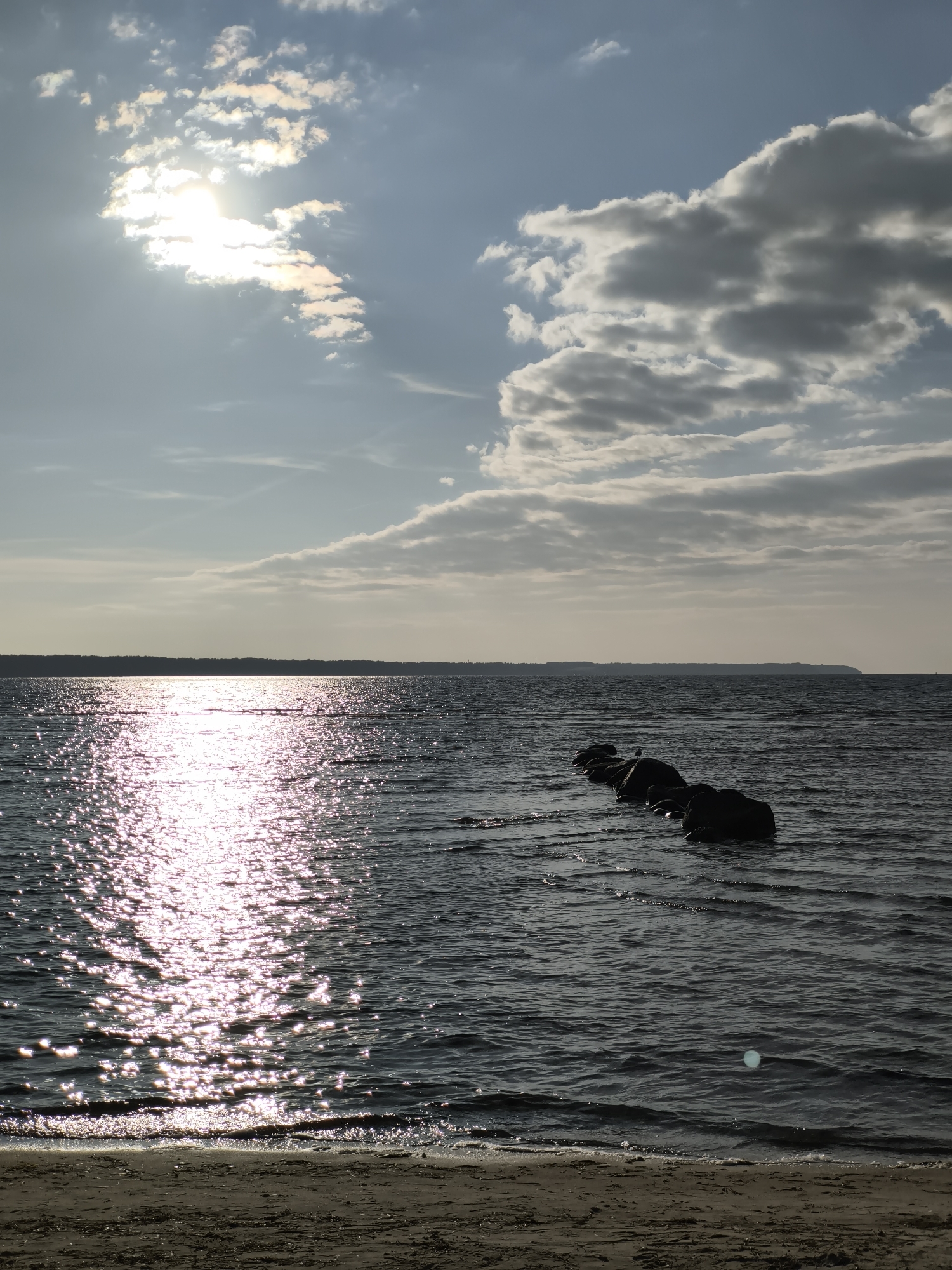 The image depicts a serene coastal scene with the sun shining brightly through scattered clouds, casting shimmering reflections on the dark water. In the foreground, a sandy shore leads to the water's edge, where gentle waves lap against the beach. A line of large, dark rocks emerges from the water, forming a breakwater. One of the rocks has a small bird perched on it. The horizon is marked by a silhouetted tree line, suggesting a forested coastline in the distance. The overall atmosphere is peaceful and natural. This scene is likely from the Tallinn Bay area in Tallinn, Estonia, given the coastal geography and general appearance.