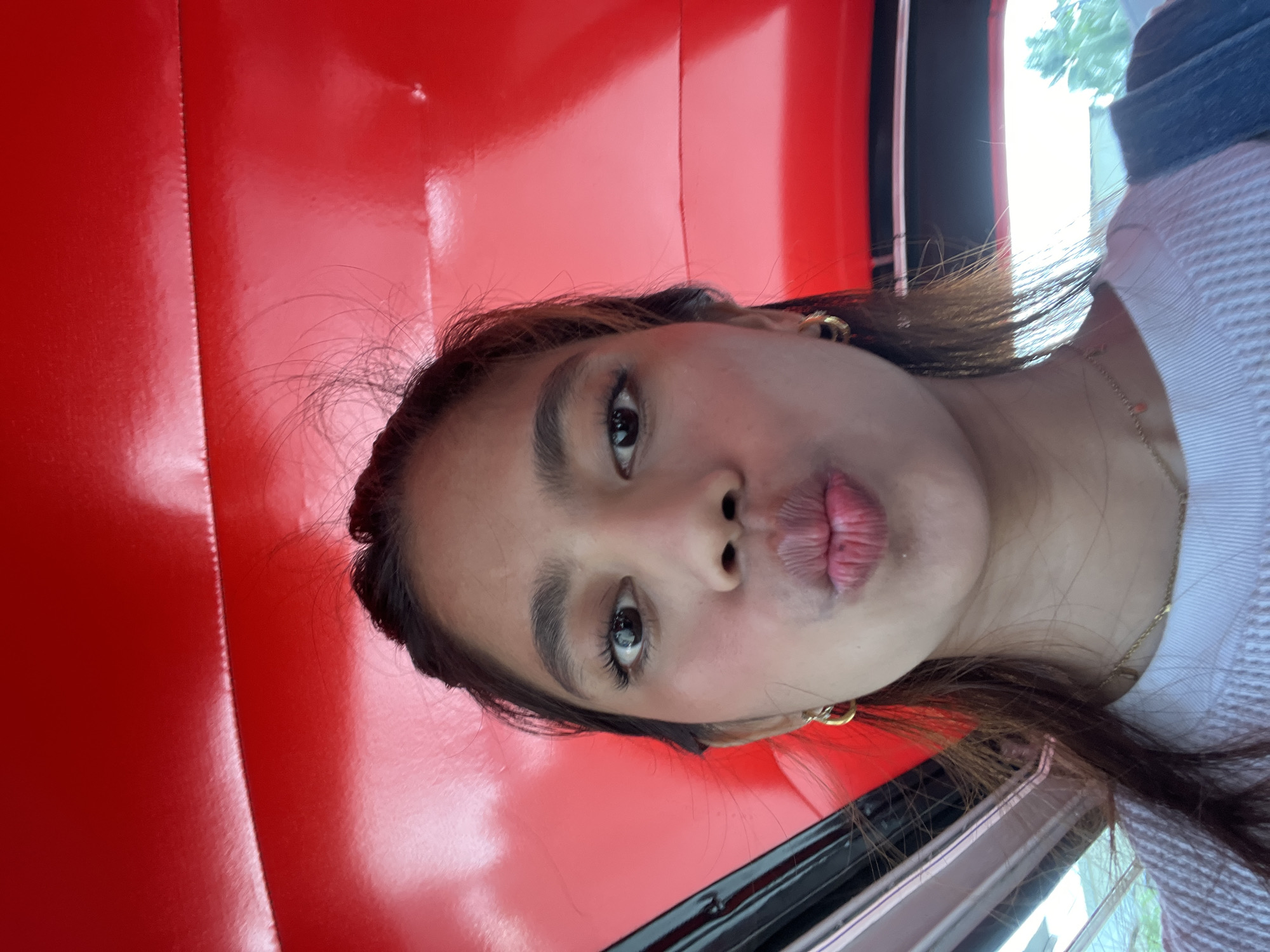 A close-up selfie of a young Filipina woman is captured inside a vehicle with a vibrant red interior. The woman is pouting her lips, with her eyes looking directly at the camera. She has brown hair with some strands falling on her face and is wearing gold hoop earrings and a gold necklace. The red interior of the vehicle is a dominant feature of the background, suggesting it might be a passenger interior of a tricycle or jeepney, common in the Philippines. The context of Panabo City, Philippines, indicates this image was taken within that urban environment. The lighting suggests it is daytime, possibly with sunlight filtering in from outside the vehicle.