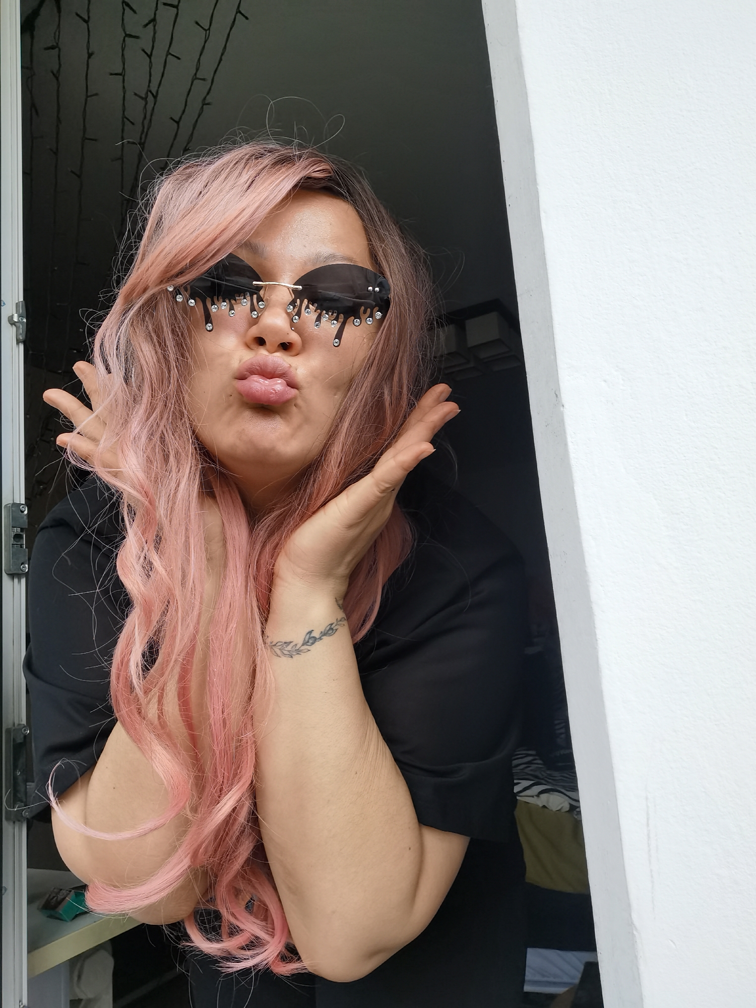 A woman with pink hair and black sunglasses adorned with dripping crystal accents stands in a doorway. She is puckering her lips and holding her hands up to her face. She is wearing a black top, and a tattoo is visible on her left wrist. Behind her, a room with hanging lights can be seen. The location is Chaikovsky, Russia.