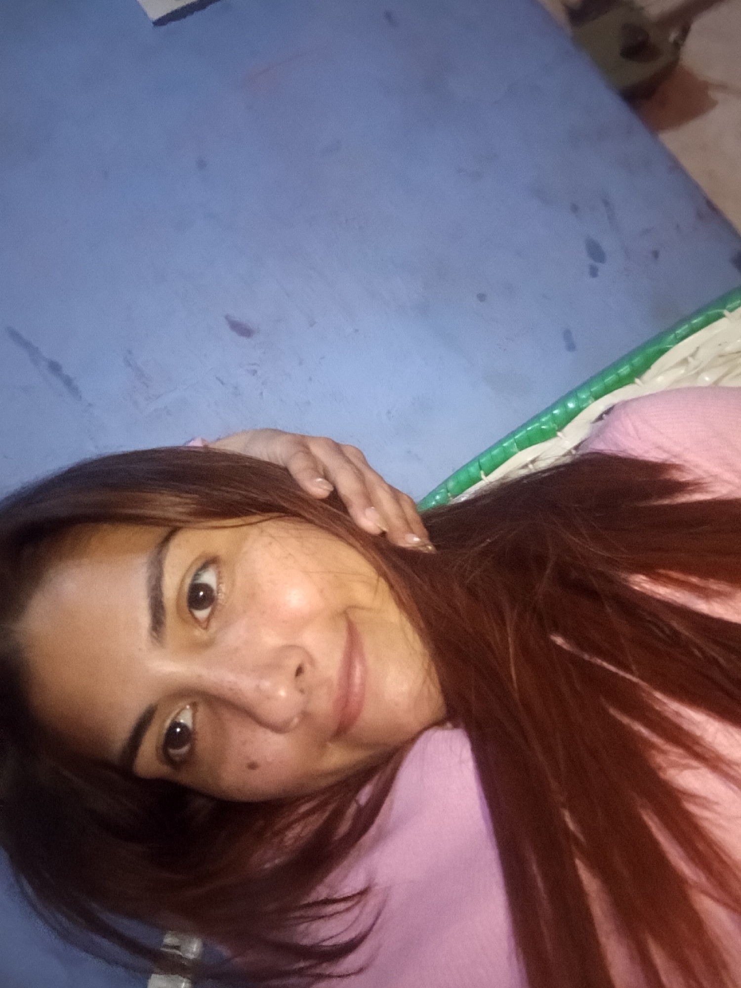 A woman with reddish-brown hair, wearing a pink shirt, is lying down. She has freckles on her face and is looking at the camera with a slight smile. Her right hand is resting on her head. She appears to be lying on a woven mat with a green and white border. The background is a light blue wall. The location is reported to be in the city of Juan Griego, Venezuela, although there is nothing visually within the image to confirm this.