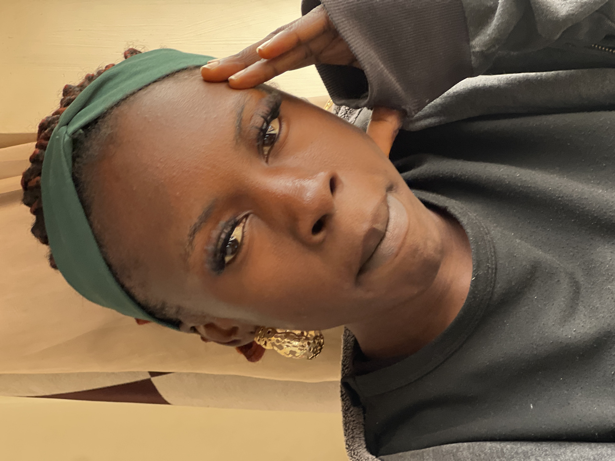 A close-up, eye-level shot shows a young woman wearing a green headband and large gold earrings. She has on a dark gray jacket over a black t-shirt and is touching her forehead with her right hand. Her hair is braided. The background consists of a light beige curtain with a brown pattern. The scene is indoors, likely in a residential setting in Jos, Nigeria.