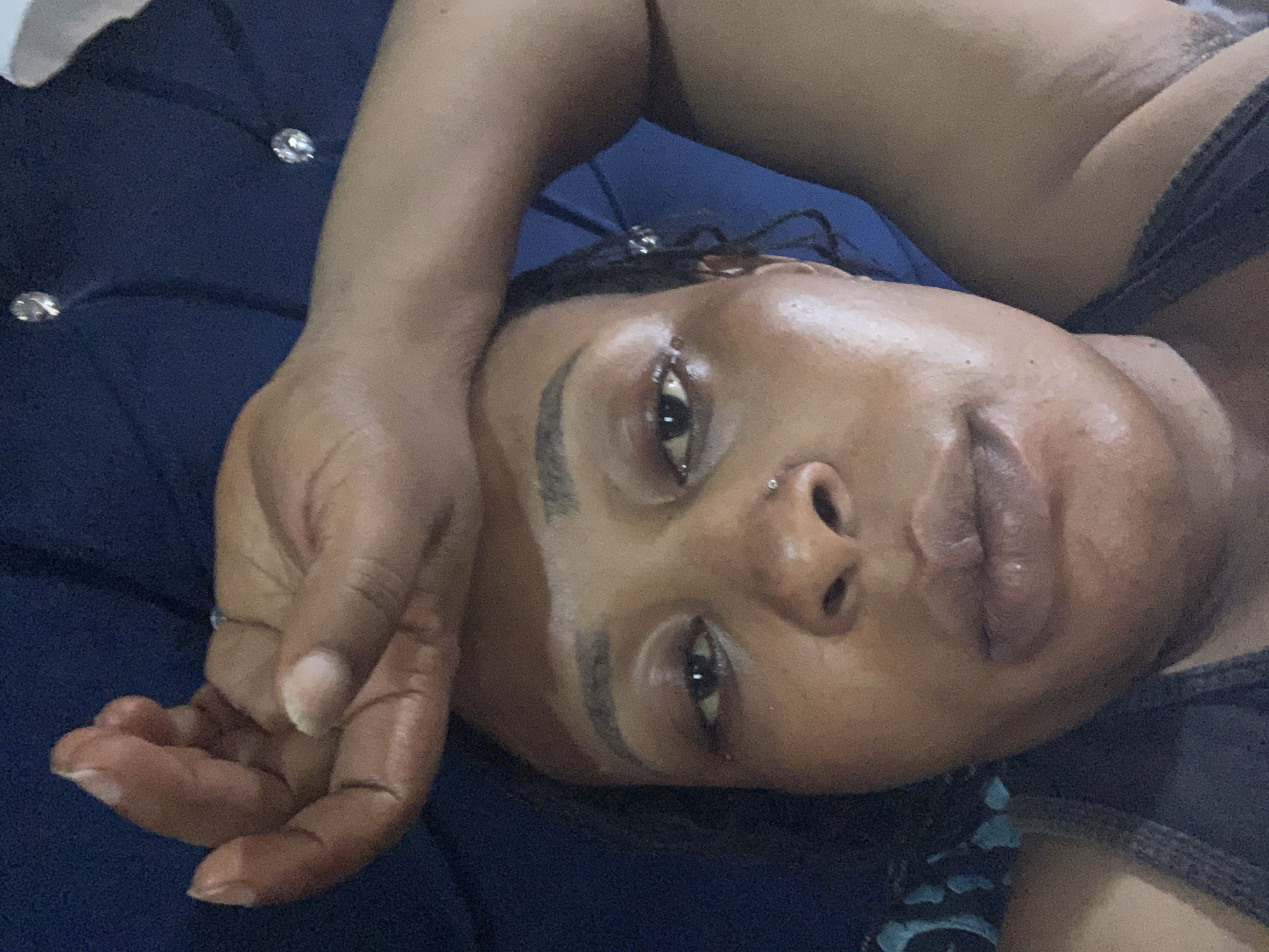 A close-up, overhead, selfie-style shot features a woman lying down, her right hand resting on her forehead, obscuring part of her face. Her eyes are partially closed, and her mouth is slightly open. She is wearing a dark gray tank top, and her dark hair is styled in braids, with some strands visible. The background is a dark blue, textured surface, possibly a bed or sofa. There are two small, clear, rhinestone-like embellishments visible on the blue surface behind her head, one above her temple and another further up. The lighting is dim, creating shadows and highlighting the sheen on her forehead and face, suggesting perspiration. The image appears to have been taken indoors. The location is stated to be Sabon Tasha, Nigeria.