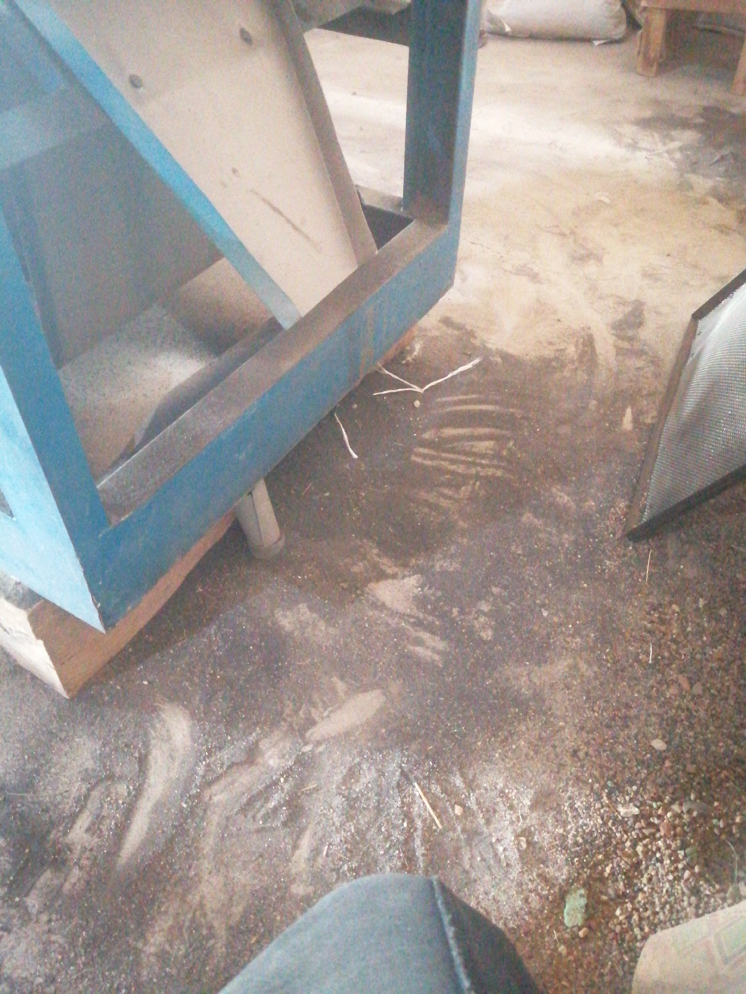 The image shows a close-up of a blue metal frame structure, possibly part of a machine or a stand, resting on a wooden plank. The structure is positioned on a concrete floor that is covered with dust and small pebbles. There are some faint handprints or streaks visible in the dust on the floor, suggesting recent activity. In the background, part of a white bag and some wooden supports are visible. To the right, a piece of mesh or screen material is angled against the floor. The overall impression is of an indoor or covered outdoor workspace, likely in an industrial or workshop setting. The presence of dust and the rough texture of the floor suggest a practical, functional environment rather than a pristine one. The location in Jos, Nigeria, indicates a likely climate where dust is a common element in such environments.