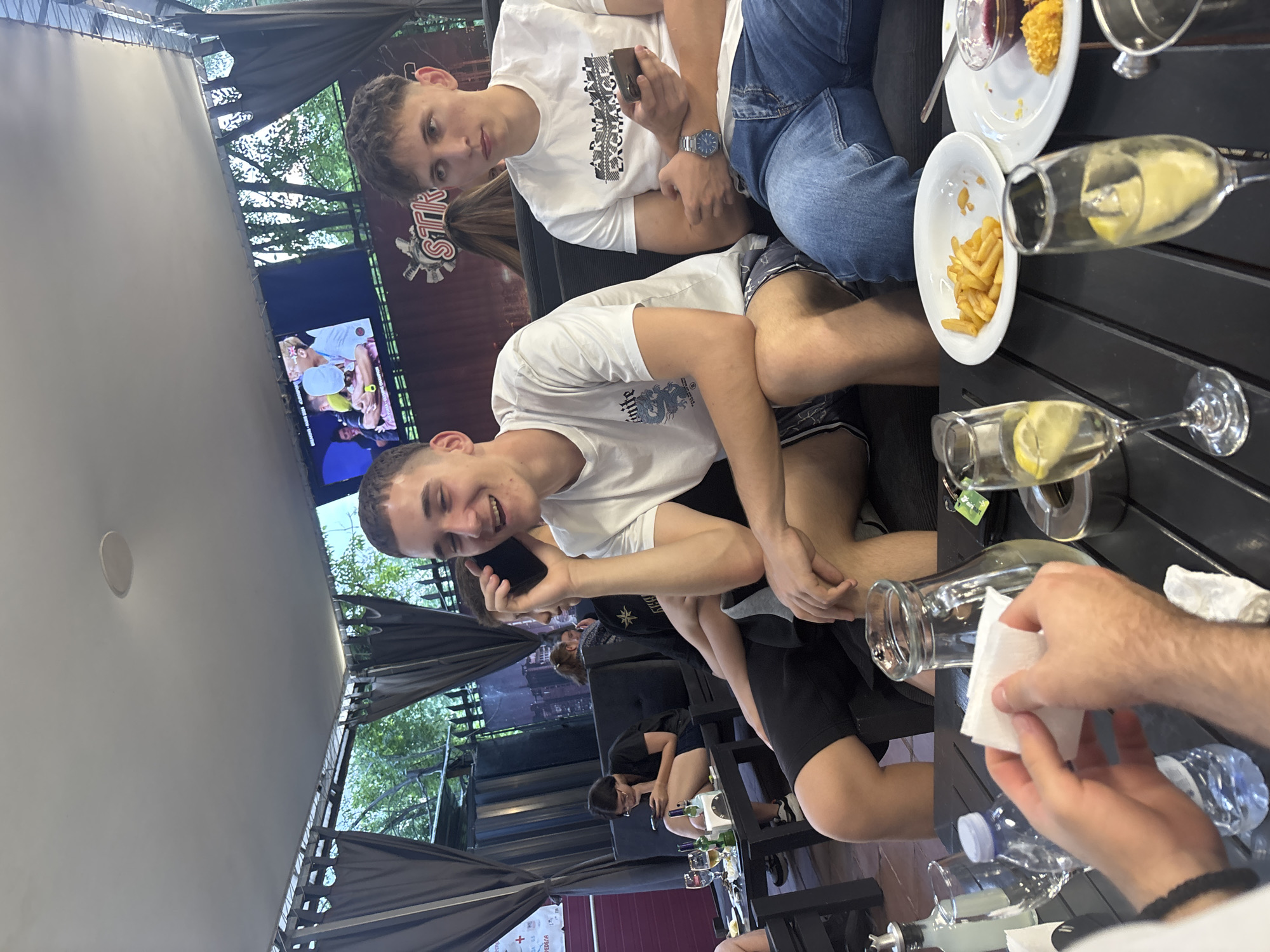 The image captures a casual social gathering at an outdoor cafe or bar identified as STIK Club in Blagoevgrad, Bulgaria. The setting features a dark-colored retractable canopy overhead and dark curtains on the sides, with natural light illuminating the scene. A back wall displays a large television screen showing a sports event, indicated by "BREAKING - PARIS 2024 - EUROSPORTS" text, and a prominent "STIK" logo. Beyond the seating area, trees and foliage are visible behind a dark fence.
In the foreground, two young men are seated on a dark sofa at a slatted black table. The man in the center, wearing a white t-shirt with a blue graphic and dark patterned shorts, is smiling widely while holding a black smartphone to his right ear, engaged in a phone call. To his right, another young man, wearing a white "ARMANI EXCHANGE" t-shirt and blue jeans, holds a black smartphone in his right hand and looks towards the left.
The table in the foreground holds various items: two champagne flutes containing a yellow liquid with lemon slices, a white plate with french fries, another white plate with a red dip and a small breaded item, a clear glass pitcher with a light-colored liquid, a clear plastic water bottle, an ashtray, and a small metal dispenser bottle. In the extreme foreground, hands are visible holding multiple white napkins.
In the mid-ground left, a woman with dark hair and glasses is seated at a separate dark table, focused on a smartphone in her hands. She wears a black t-shirt and dark shorts. Her table has two beer bottles and two glasses. The back of another person's head with light brown hair is partially visible between the two foreground men.
