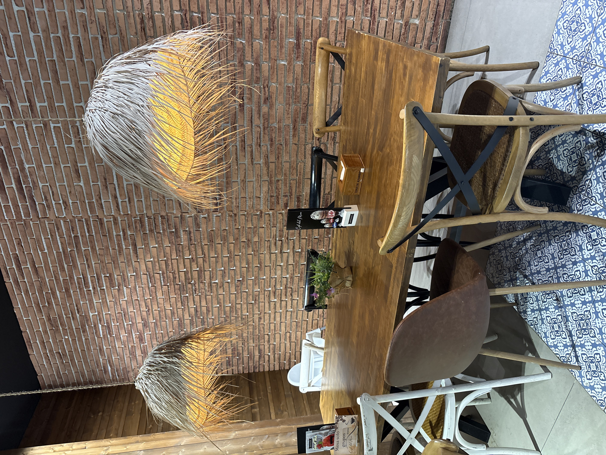 This media file captures an indoor dining setting, likely a restaurant or cafe, with a rustic and modern aesthetic. The scene features a wooden table surrounded by various chairs, including a brown upholstered one, a light-colored wooden one with a cross-back design, and a white metal chair. A brick wall forms the backdrop, adding to the textured and warm ambiance. Two large, pendant light fixtures with a natural, fibrous material hang from the ceiling, emitting a warm yellow glow that illuminates the space.
On the table, several items are visible, including a small plant in a woven pot, a small wooden box, and a menu. One menu, in particular, displays text that appears to be in Spanish, with phrases like "Cafe del Mar" and "Mesa solo grupos." The table itself is made of polished wood, reflecting the light.
The overall impression is that of a cozy and inviting dining environment. The lighting suggests it might be evening or a dimly lit interior. The presence of the chairs around the table implies that this is a space designed for patrons to sit and dine. No people are visible in the image, so no specific activity is depicted, but the setup strongly suggests a dining or gathering context. The location is identified as Valencia, Spain, which could inform the style of the decor or menu if further context were available. The blue and white patterned fabric visible on the right side of the image may be part of the seating or table covering, adding a touch of decorative flair to the scene.