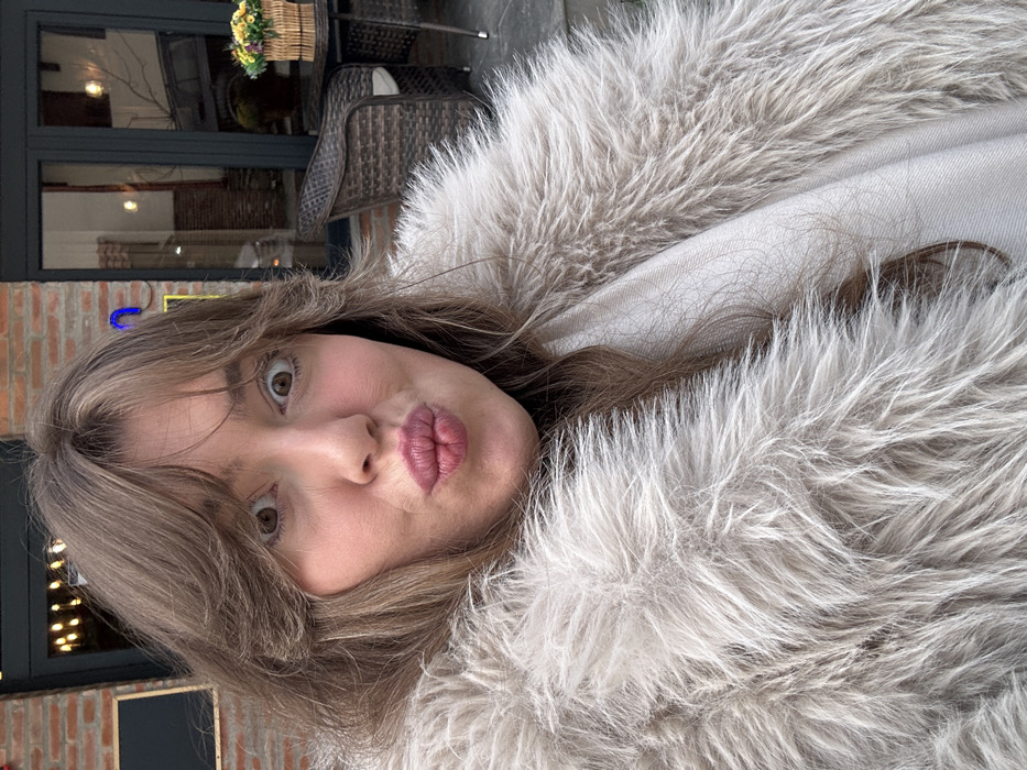 A light brown-haired female subject is depicted in a close-up, exhibiting a wide-eyed expression with puckered lips. She is dressed in a light grey shaggy faux fur coat, layered over a cream-colored garment. The background indicates an outdoor cafe environment located in Sighnaghi, Georgia. Visible elements include a dark wicker chair with a light cushion and a round, dark-colored table. A wicker basket containing yellow and purple artificial flowers rests on the table. Behind this setup, a brick building facade with a dark-framed glass door reflecting interior lights is present. The overall lighting is subdued.
