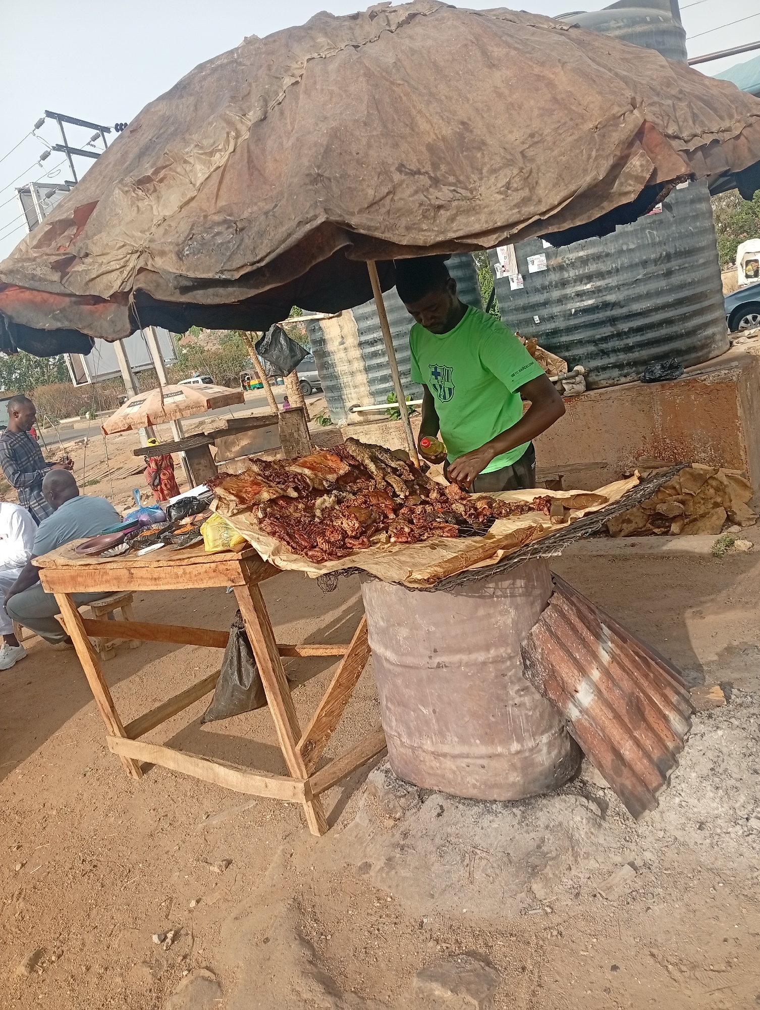 The image captures a roadside food vending scene in Jos, Nigeria. An individual wearing a green t-shirt is seen working at a makeshift food stand. A large pile of what appears to be grilled meat sits on a surface covered with brown paper. The table is supported by a metal drum. An umbrella provides shade for the stand. Customers are gathered at the wooden table to the left of the food stand. Behind the stand is a concrete structure and two water tanks. Cars, trees, and buildings can be seen in the background. The ground is dusty.